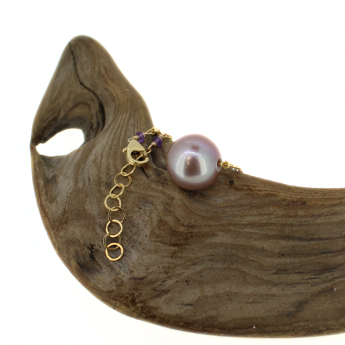 A gold chain necklace with a single lustrous purple pearl and small purple beads, displayed on a curved piece of driftwood against a white background.