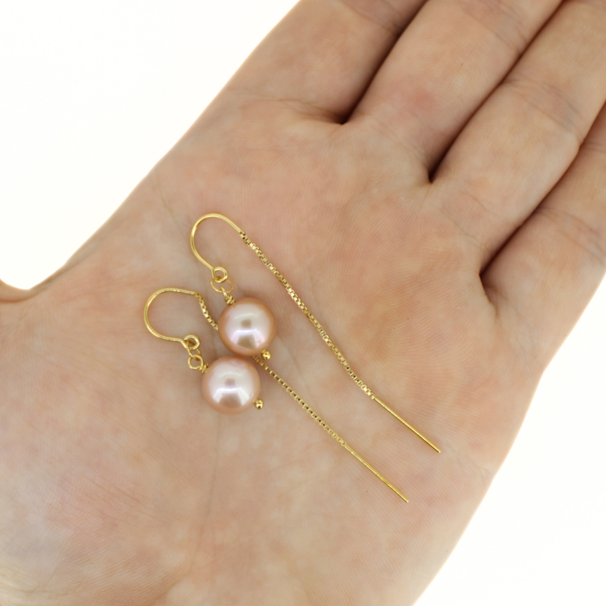A pair of gold threader style earrings with round pinkish pearl-like beads and long dangling chain that hangs behind the ear, displayed on an open hand against a white background.