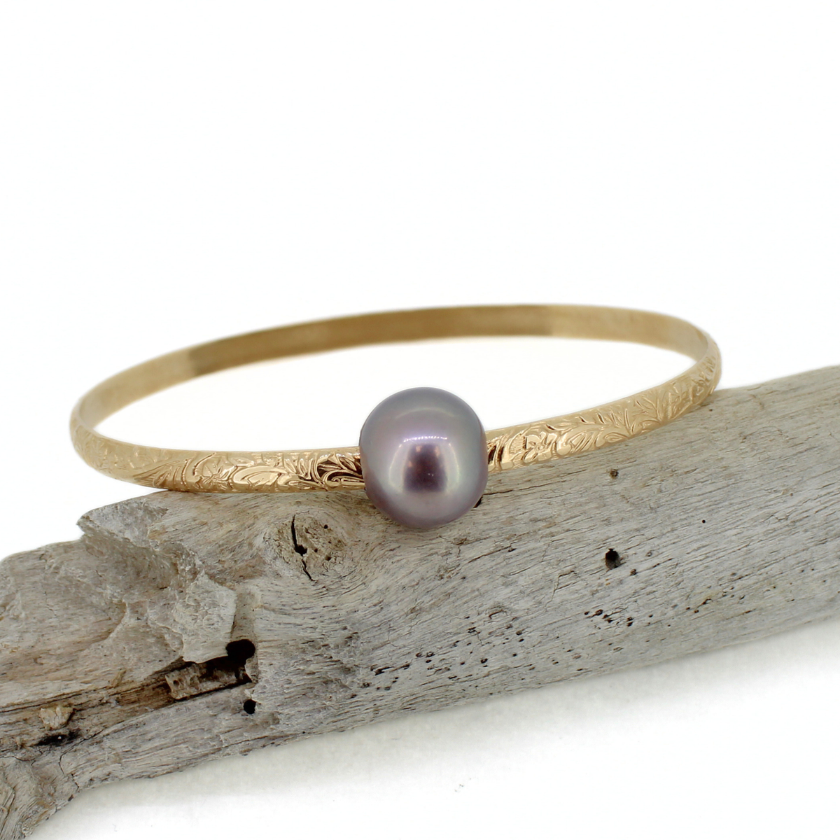 A delicate gold bangle bracelet with a single iridescent purple pearl, resting on a piece of weathered driftwood against a white background.