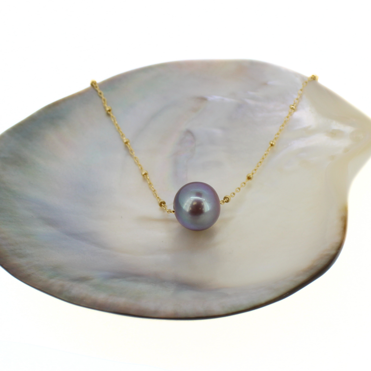 A gold chain necklace with a single round purple pearl pendant, displayed on a white mother-of-pearl shell against a white background.