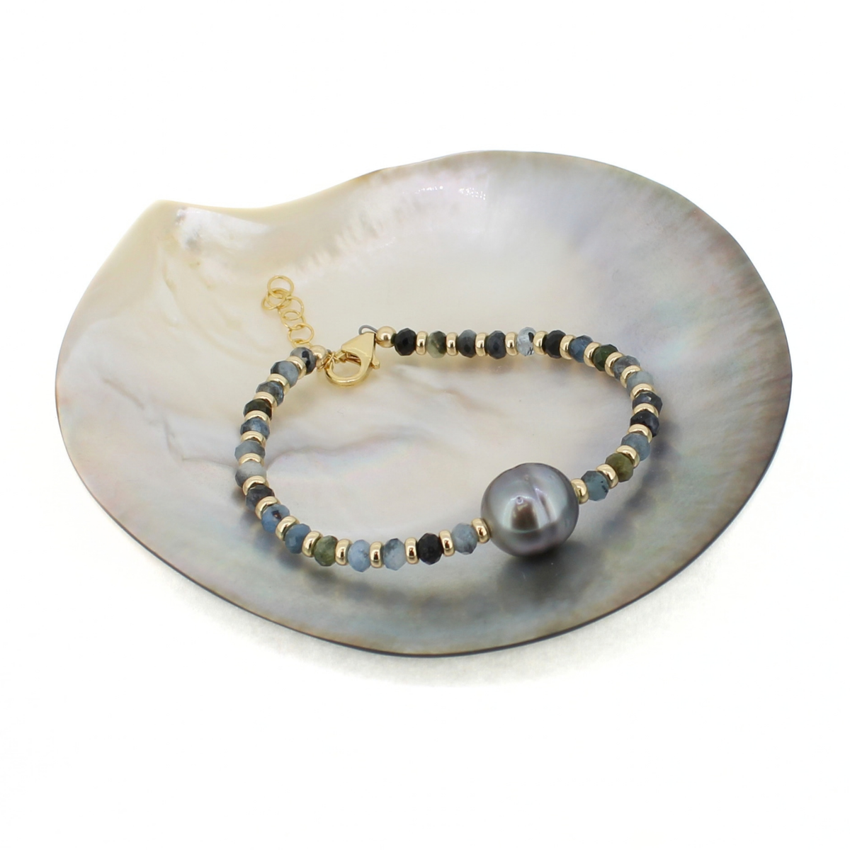 A beaded bracelet with a large dark gray pearl and gold clasp, displayed on a white shell dish against a white background.