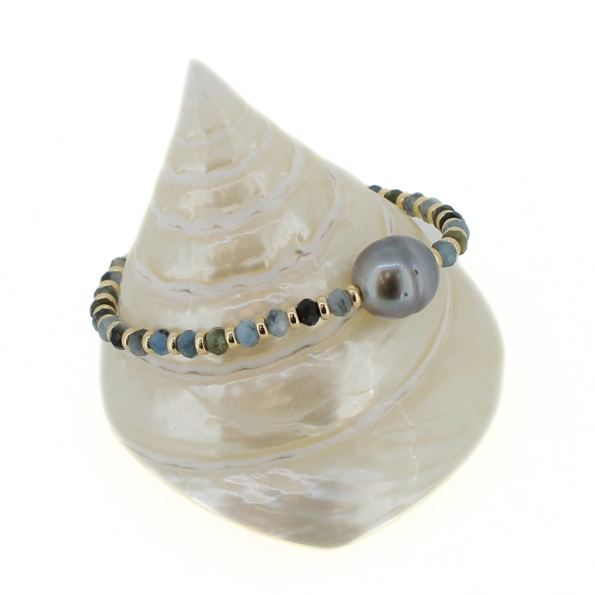 A beaded bracelet with a large gray pearl and small blue and green beads, displayed on a white seashell stand against a white background.