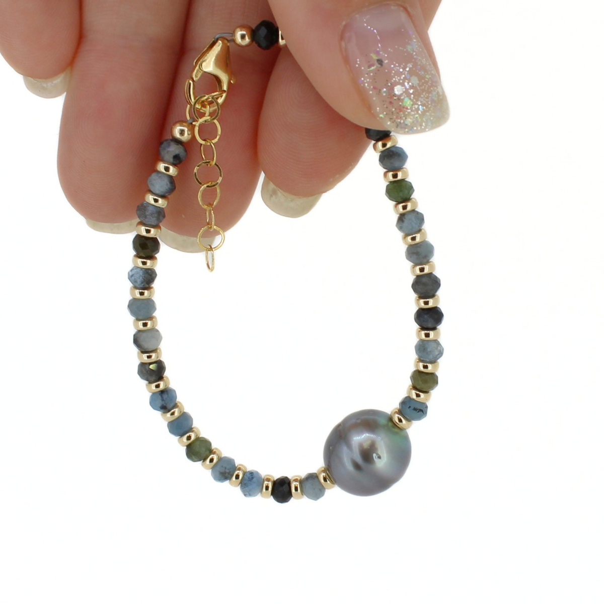 A delicate beaded bracelet with a large iridescent pearl, gold spacer beads, and small blue and green beads, held by a hand with glittery nail polish against a white background.