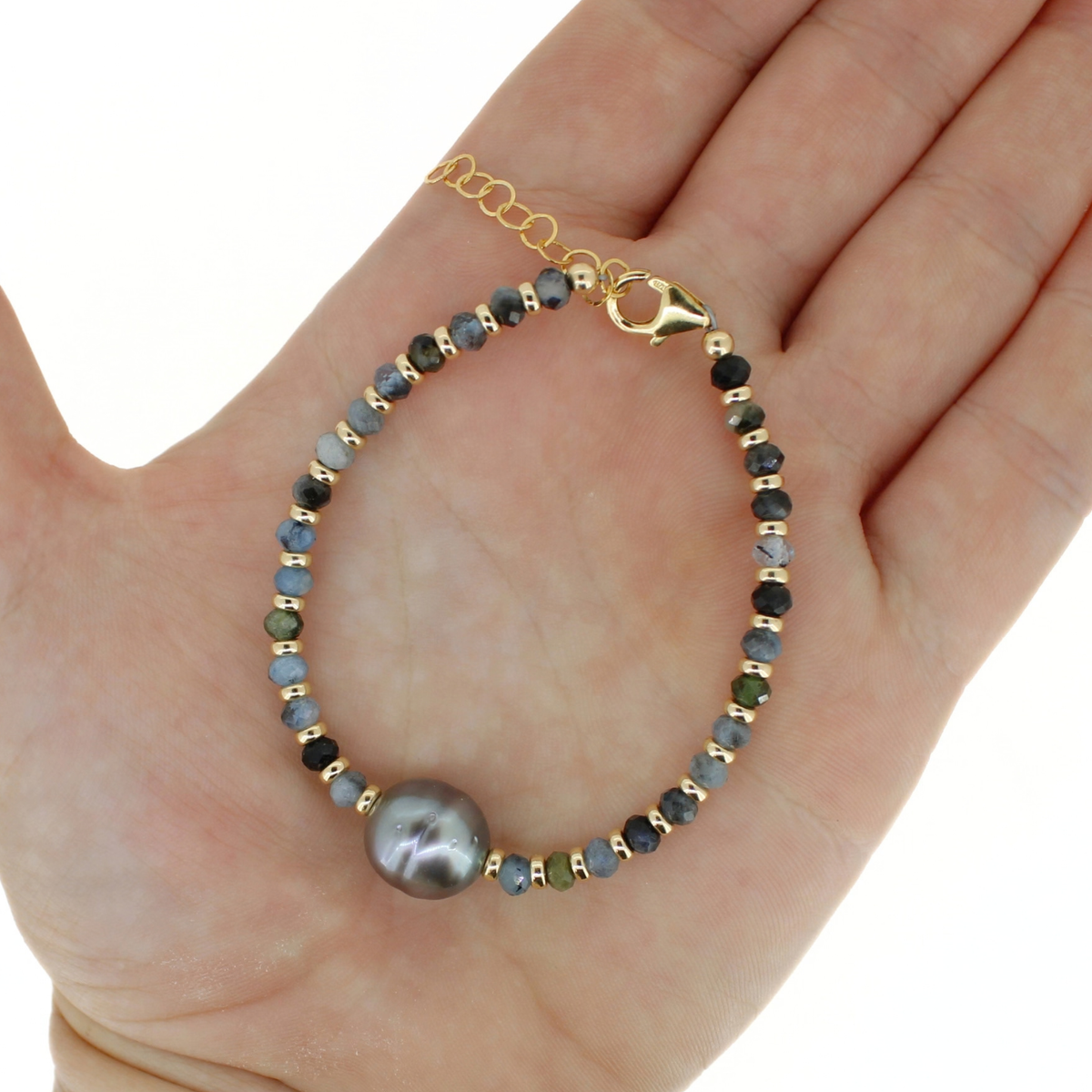 A gold bracelet with a large gray pearl and small multicolored beads, displayed on an open hand against a white background.