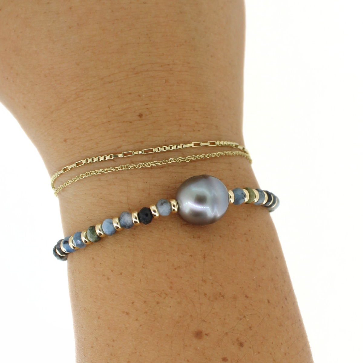 A close-up of a person's wrist wearing two delicate bracelets: a thin gold chain bracelet and a beaded bracelet featuring a large gray pearl and small blue, green, and gold beads.