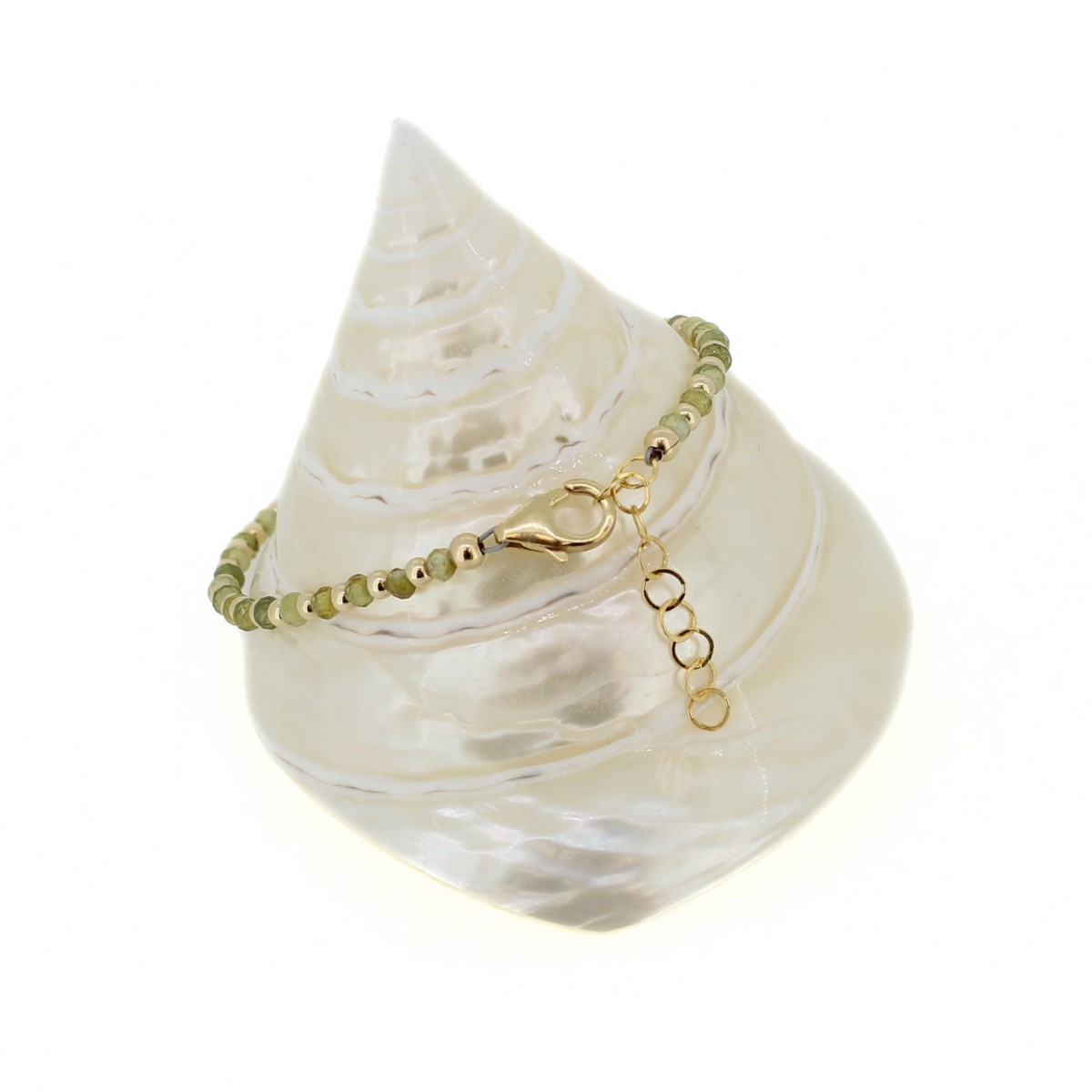 A delicate beaded bracelet with small green beads and gold metal accents, displayed on a white seashell stand against a white background.