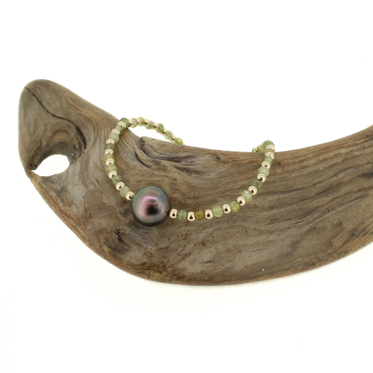 A delicate beaded bracelet with a large iridescent black pearl, displayed on a curved piece of driftwood against a white background.