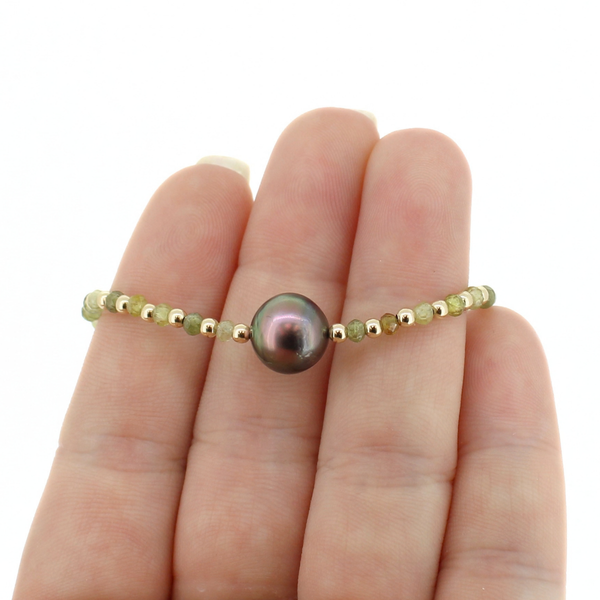 A delicate beaded bracelet with a central dark iridescent pearl, displayed on a person's fingers against a plain white background.
