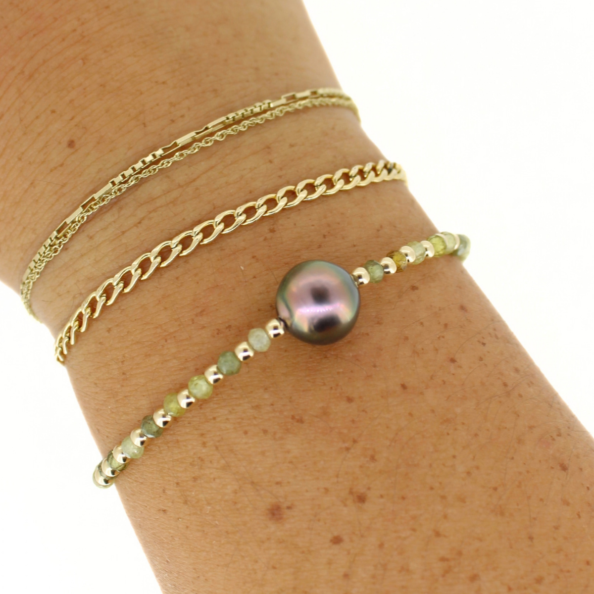 A close-up of a person's wrist wearing three layered gold bracelets: a fine Figaro chain, a curb chain, and a beaded bracelet featuring a round iridescent dark pearl and small green beads.