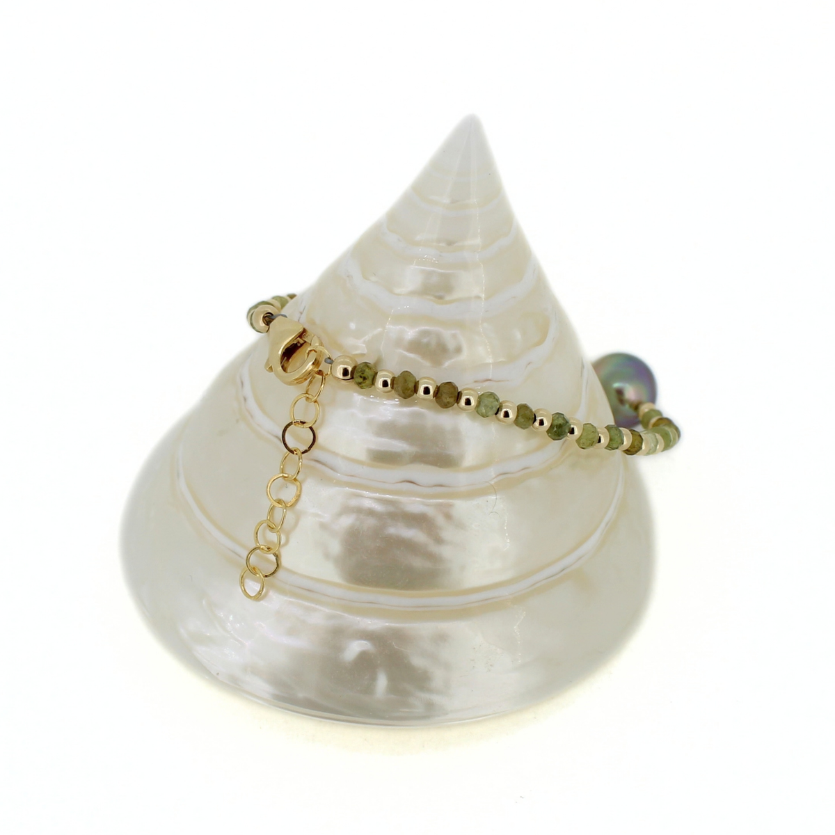 A delicate beaded bracelet with small green beads and gold spacer beads, featuring a gold clasp and extender chain, displayed on a white seashell jewelry stand against a white background.