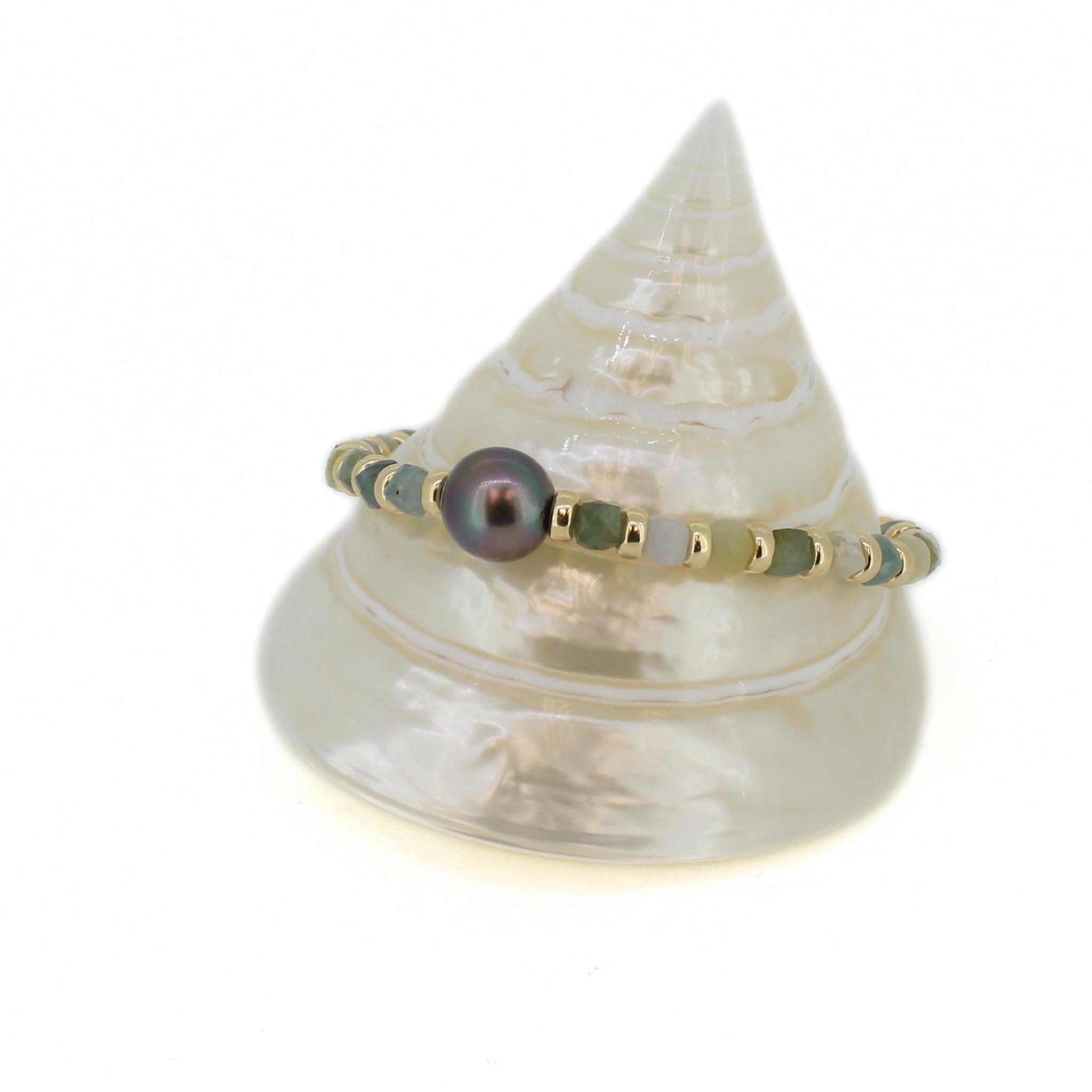 A delicate beaded bracelet with a central iridescent dark pearl and green jade beads, displayed on a white seashell stand against a white background.