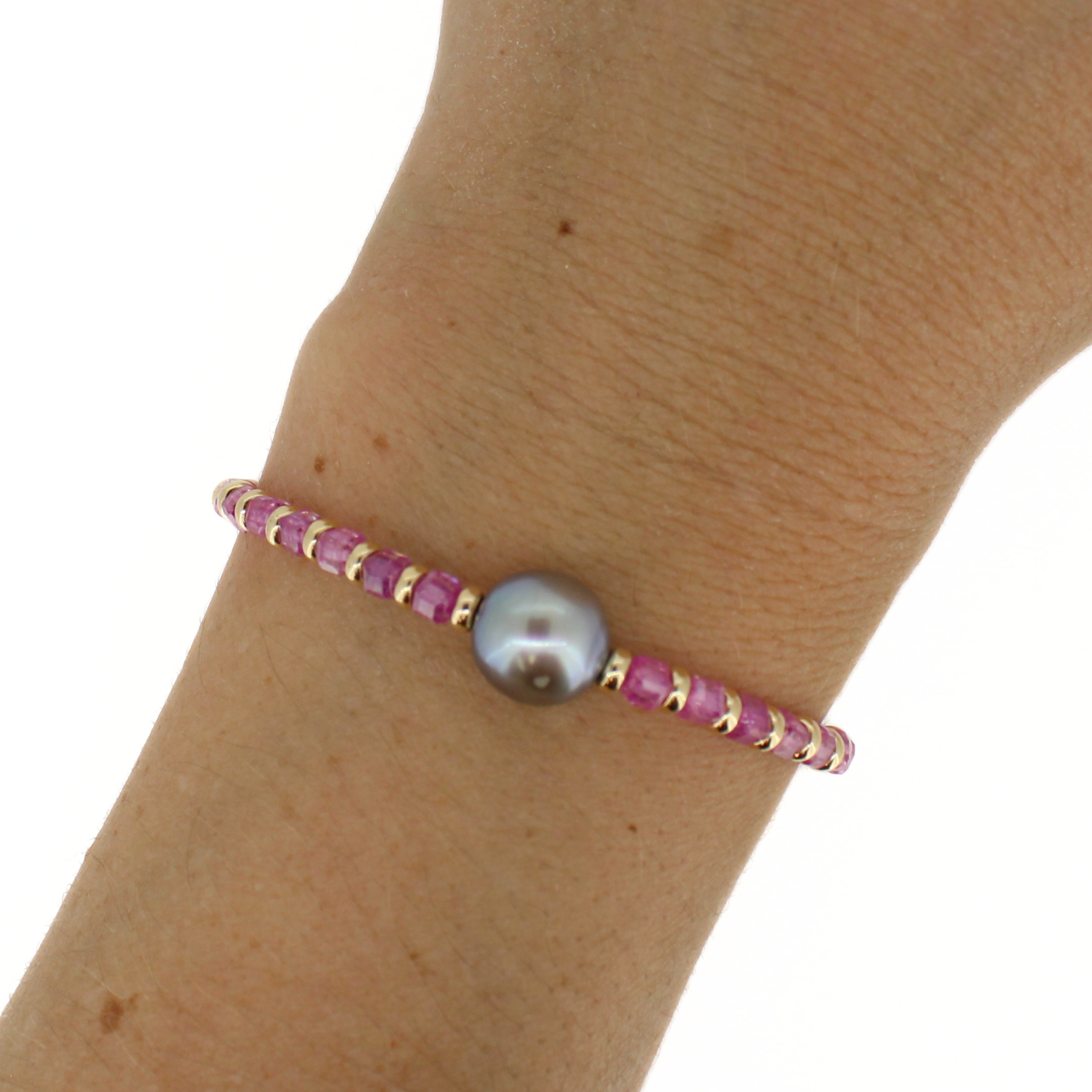 A close-up of a person's wrist wearing a delicate bracelet made of pink faceted beads, gold spacer rings, and a central round gray pearl.