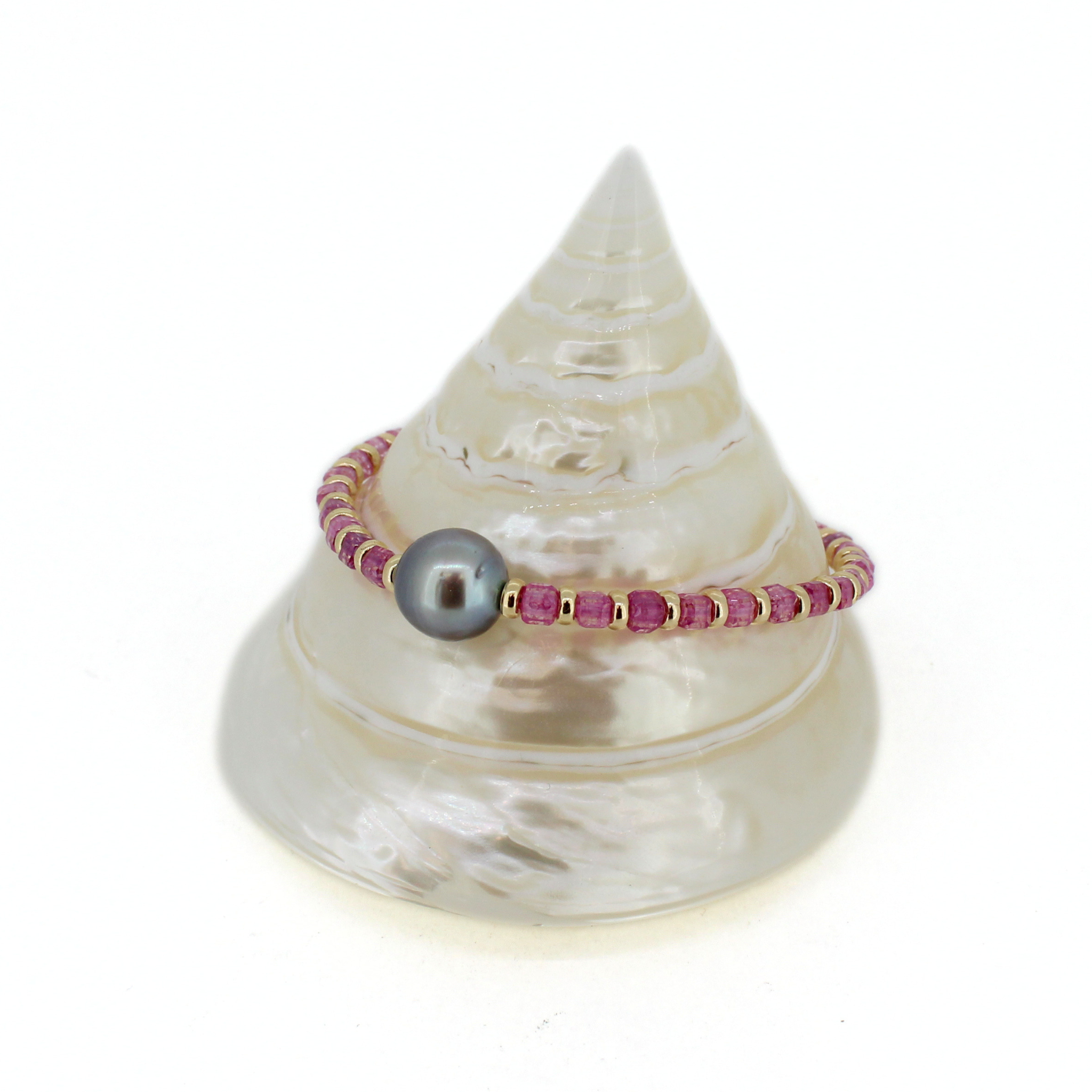 A beaded bracelet with pink faceted beads, small gold spacer rings, and a single Tahitian pearl, displayed on a white seashell stand against a white background.