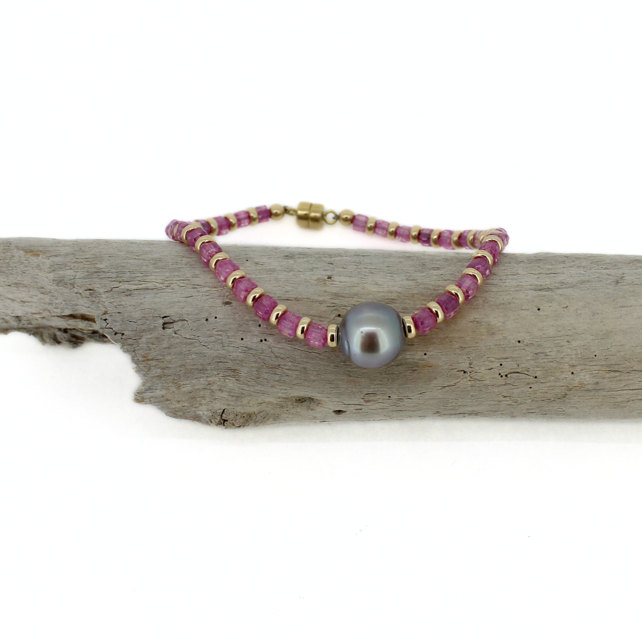 A beaded bracelet with pink cube-shaped topaz beads, small gold spacer beads, and a central gray Tahitian pearl, displayed on a piece of weathered driftwood against a white background.