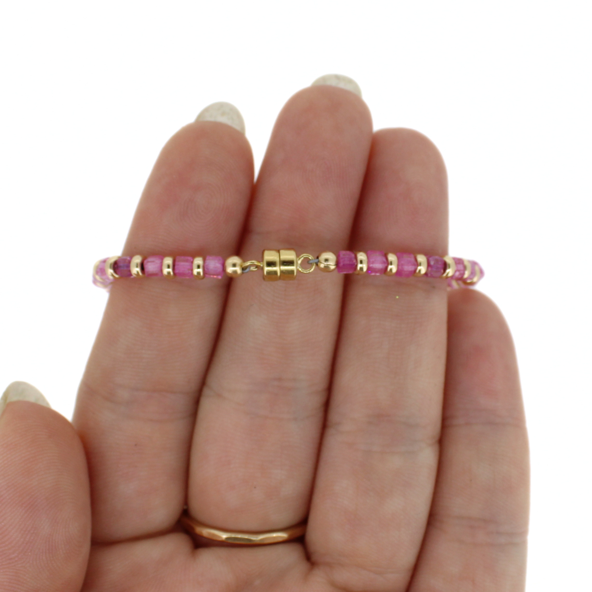 A delicate beaded bracelet with pink faceted beads and gold spacer rings, finished off with a magnetic clasp held on a person's fingers against a plain white background.