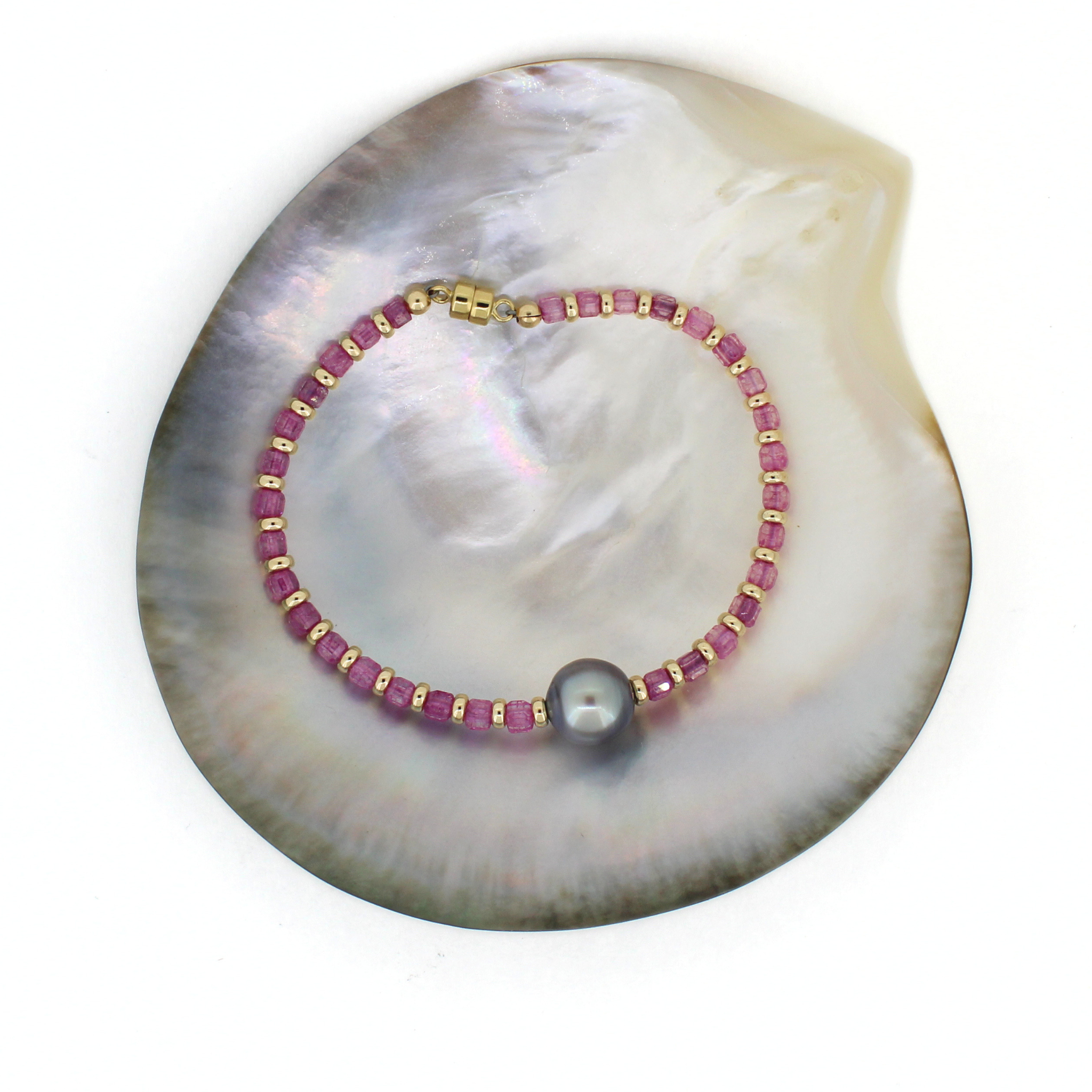 A beaded bracelet with pink faceted topaz beads, gold spacer beads, and a single gray Tahitian pearl, displayed on a white background over a mother-of-pearl shell.
