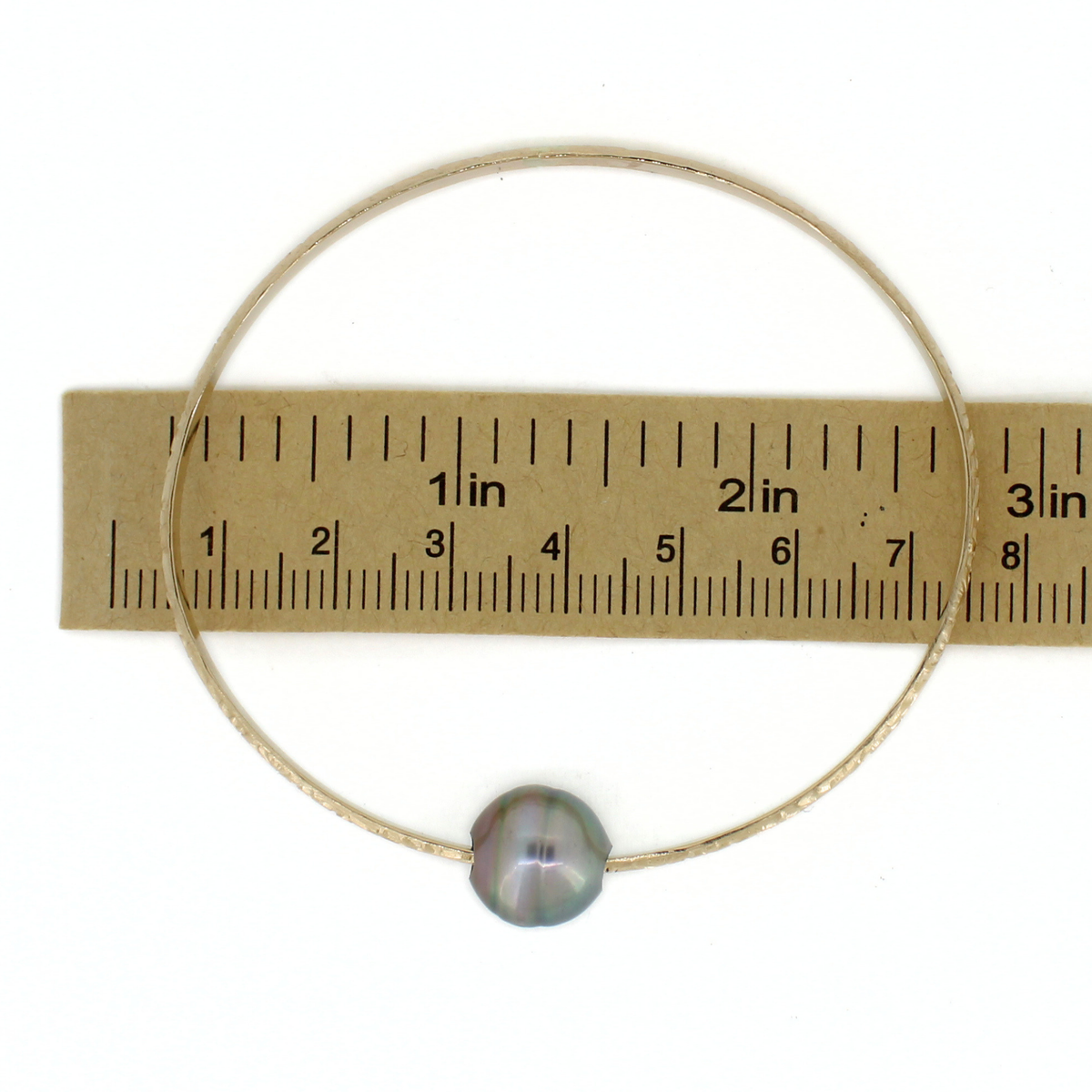 A thin gold bangle bracelet with a single round iridescent pearl bead, shown against a white background with a brown paper ruler for scale.