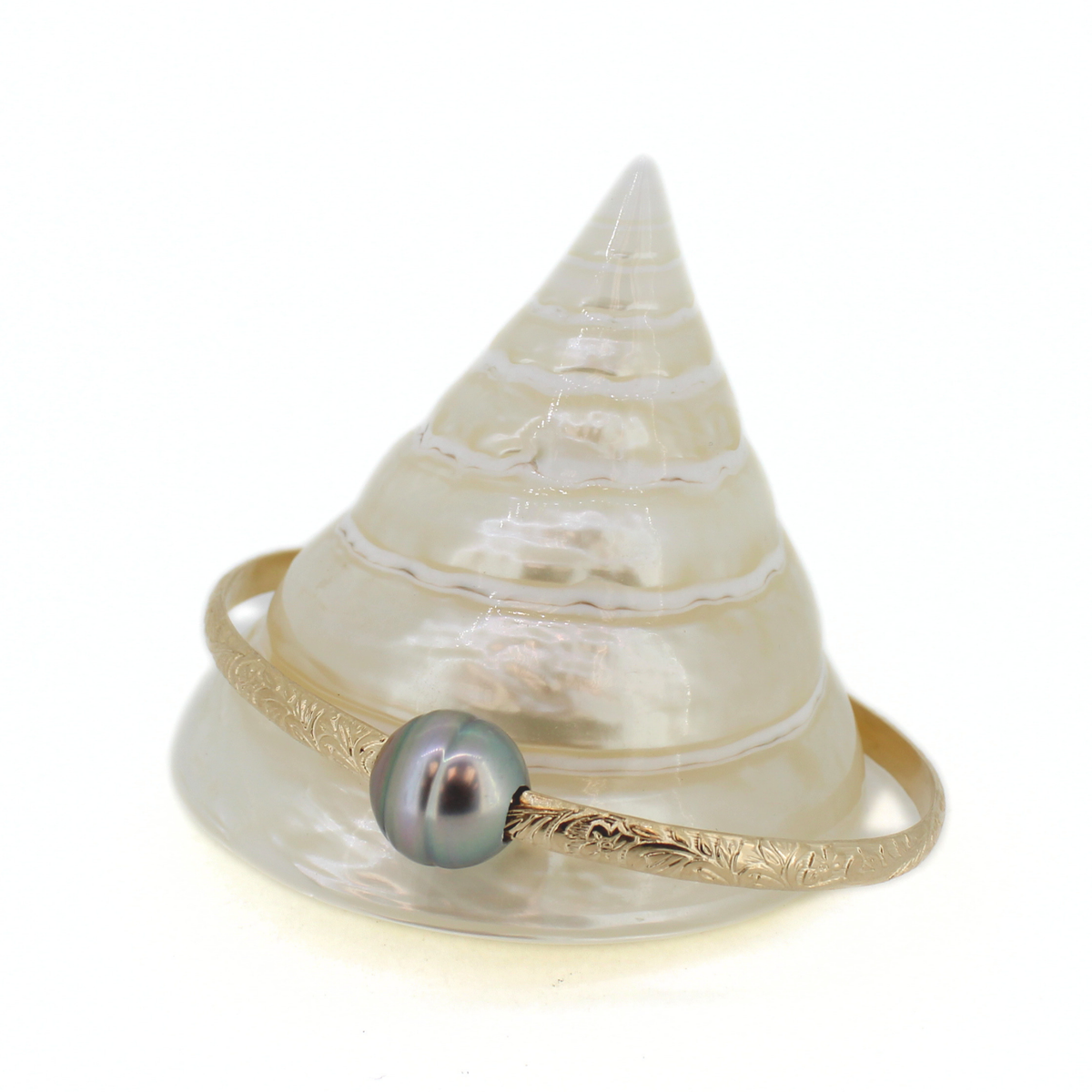 A gold bangle bracelet with a textured engraved band and a single round gray pearl, displayed on a white conical shell stand against a white background.