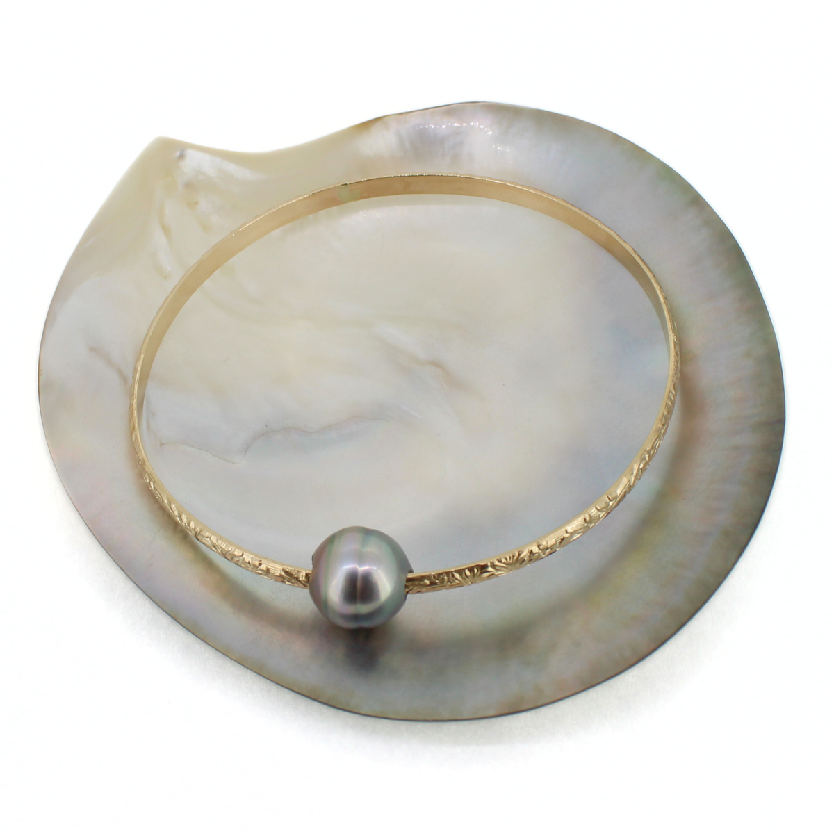A delicate gold bangle bracelet with a textured band and a single dark gray pearl, displayed on a white background over a mother-of-pearl shell prop.