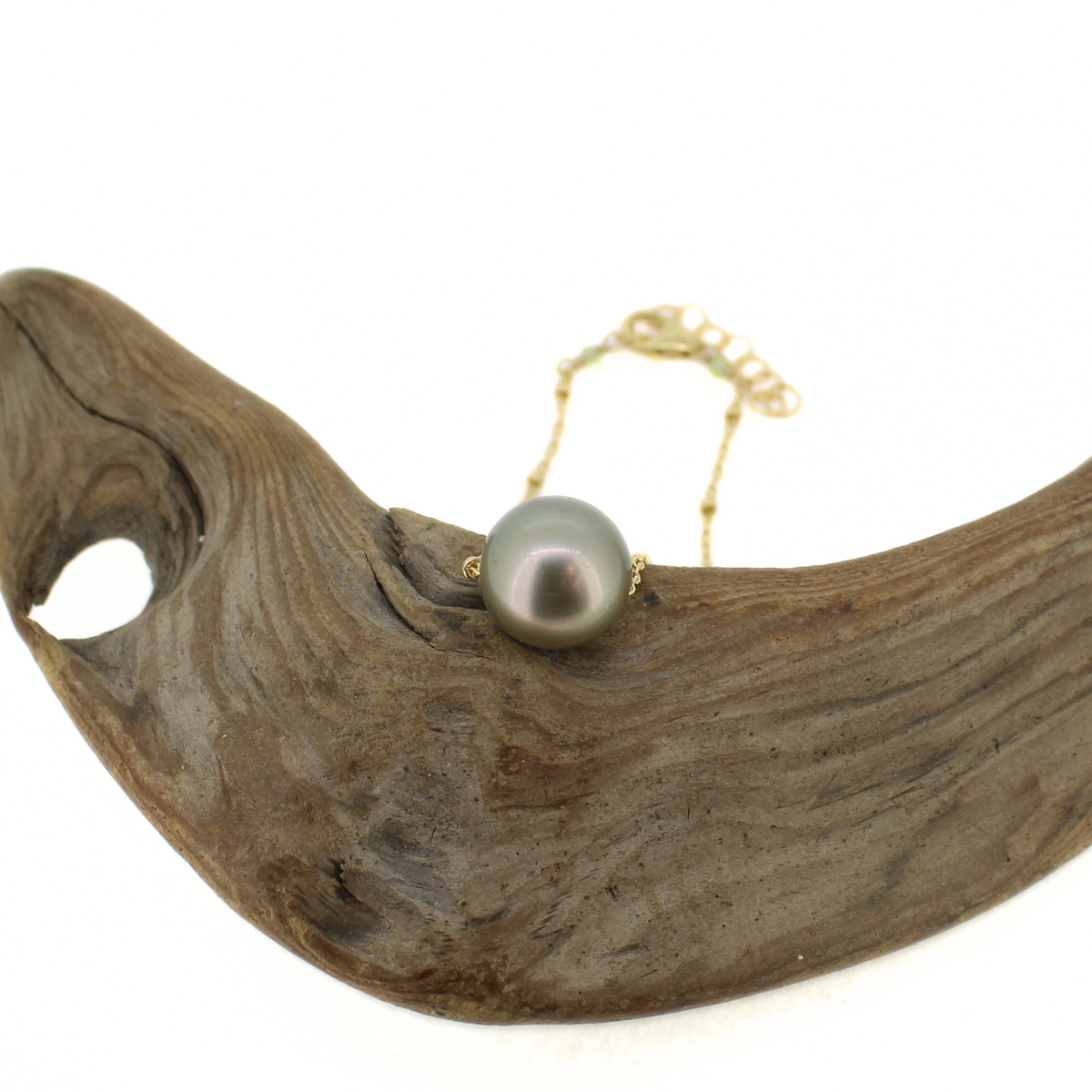 A green Tahitian pearl that floats along a gold satellite chain necklace, shown on a piece of driftwood.