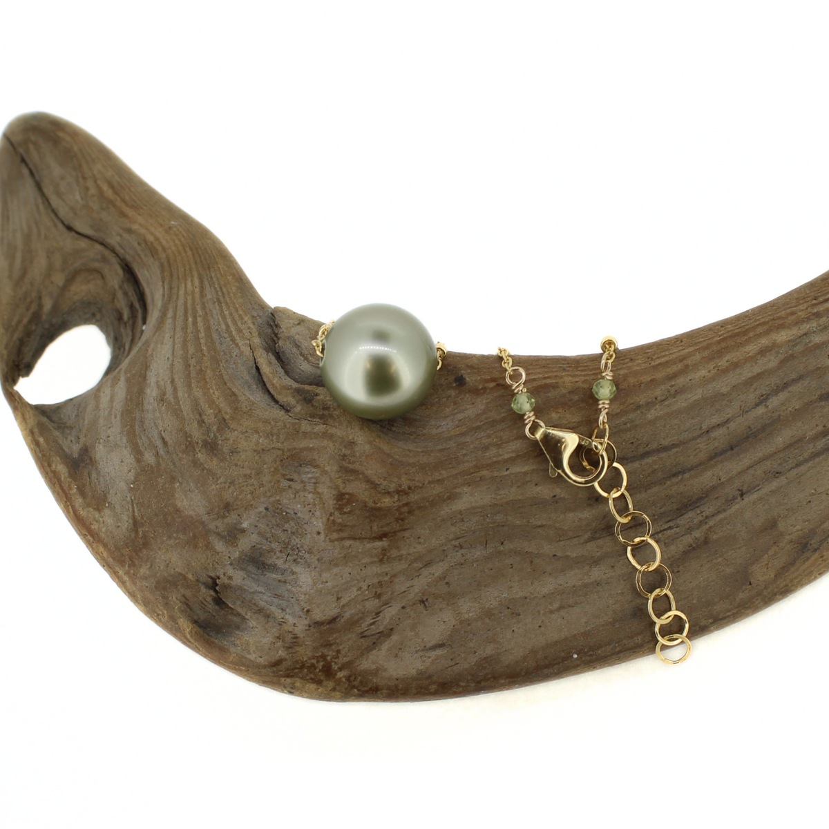 A gold chain necklace with a single large gray-green pearl and small green beads, draped over a curved piece of driftwood on a white background.