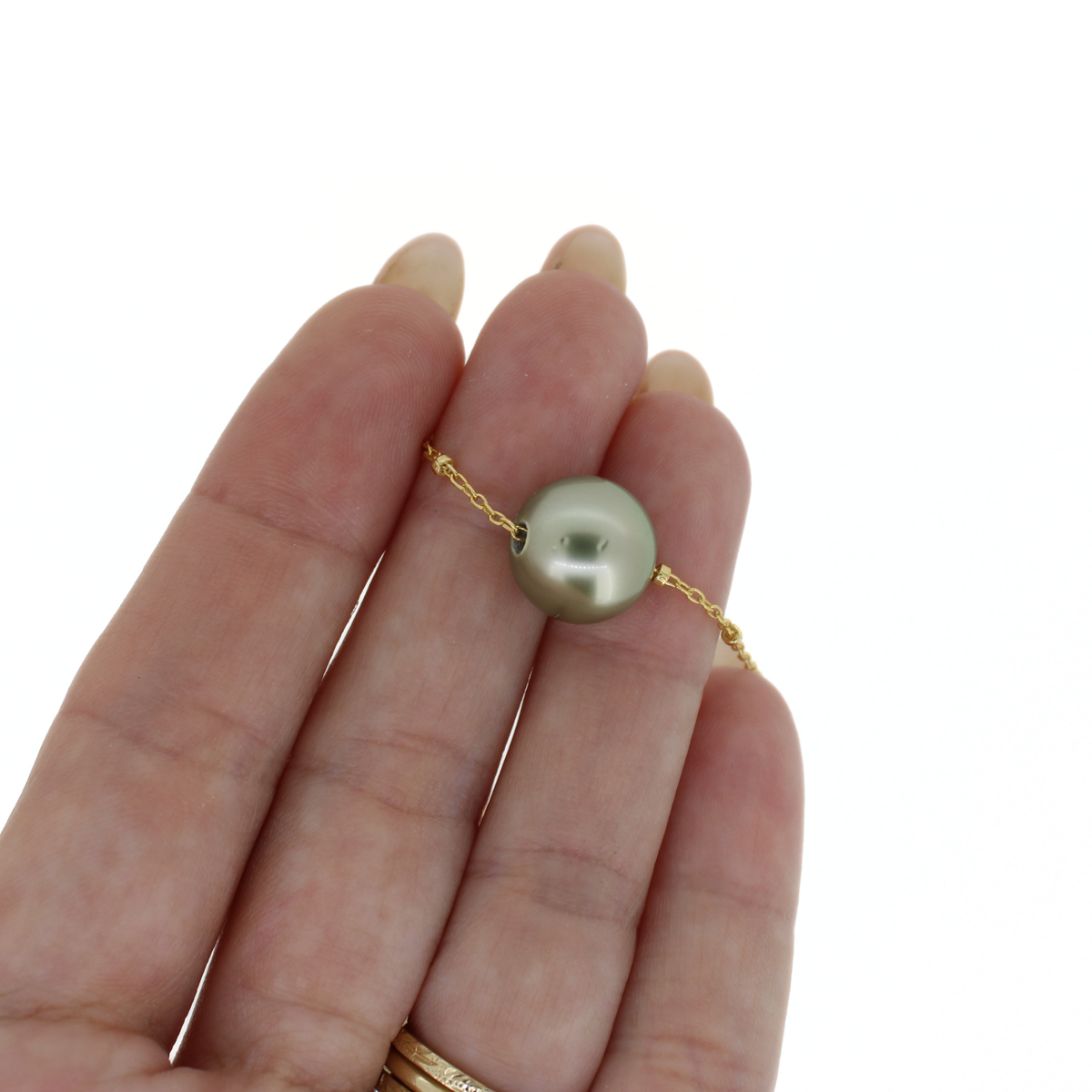 A close-up of a hand holding a delicate gold chain necklace with a single round Tahitian pearl pendant. The pearl is a light gray-green color, and the chain is gold.