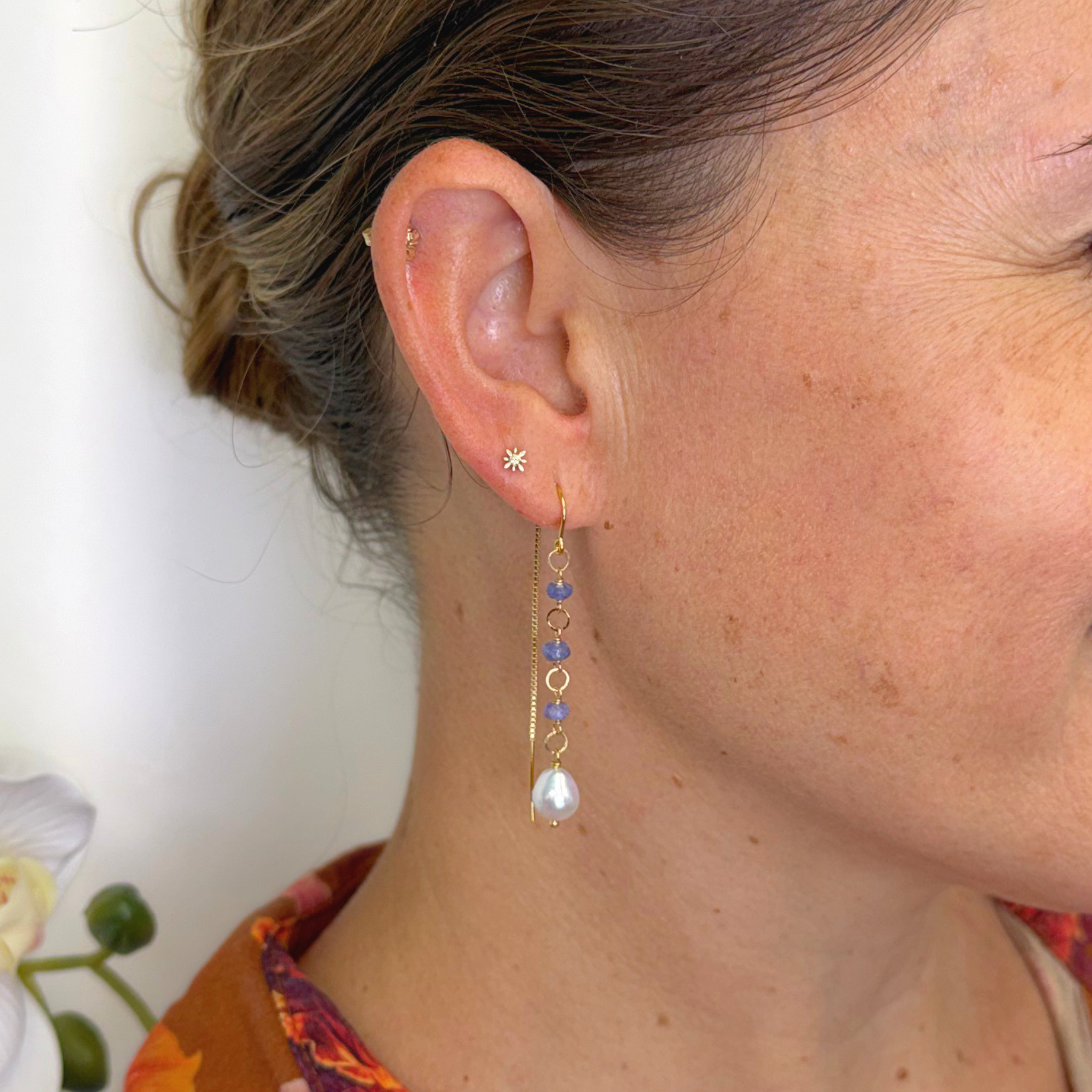 A close-up of a woman's ear wearing a delicate gold dangle earring with a small star stud, blue beads, and a teardrop pearl.