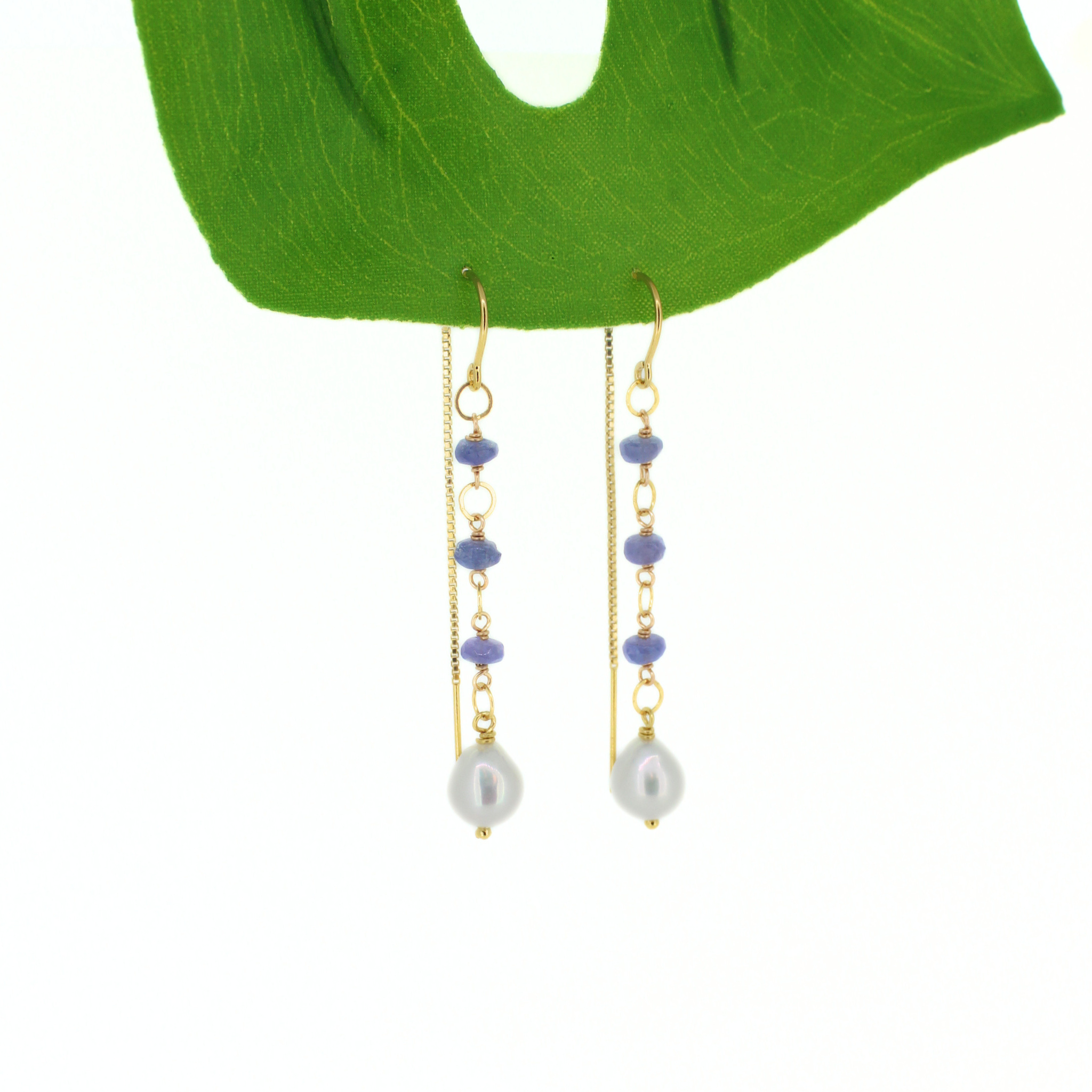 A pair of gold dangle earrings with small lavender beads and white baroque pearls, displayed on a green leaf against a white background.