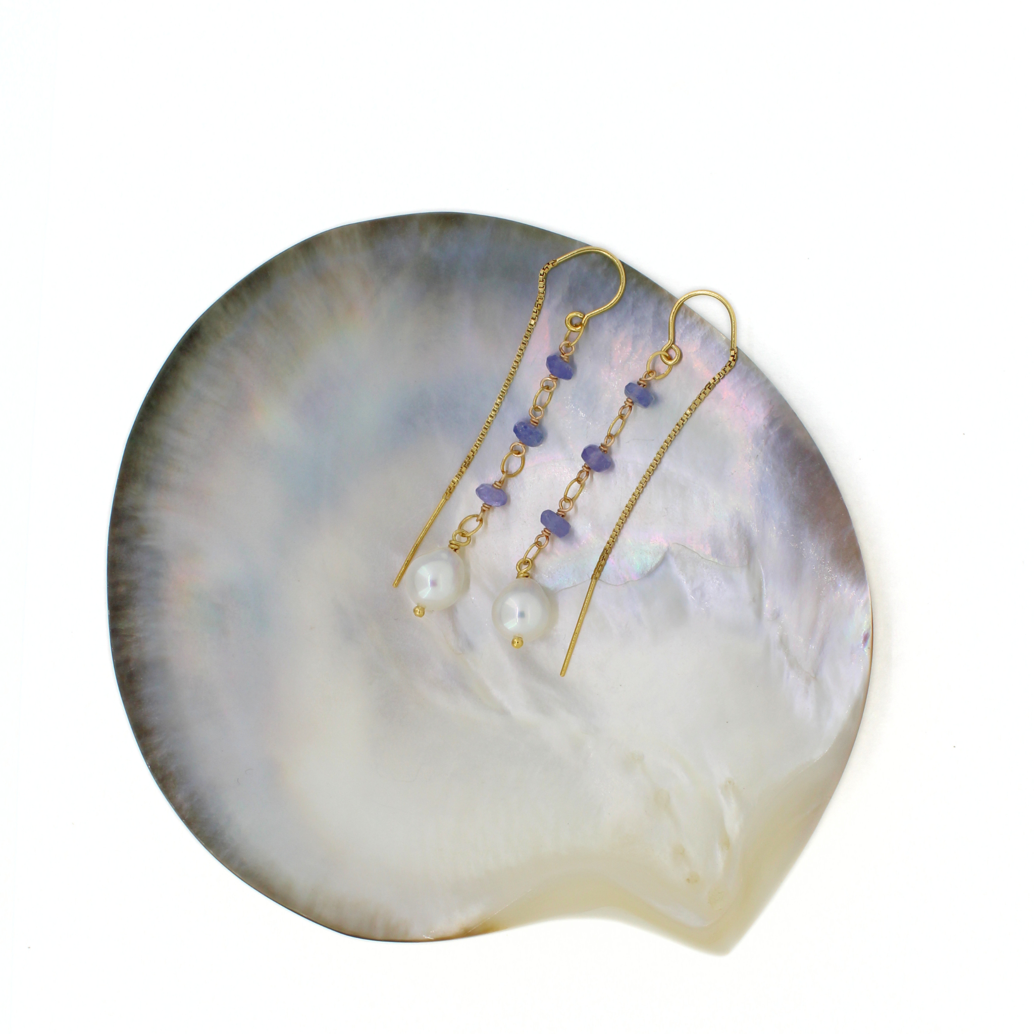 A pair of gold drop earrings with small purple beads and white pearl-like drops, displayed on a large iridescent shell against a white background.