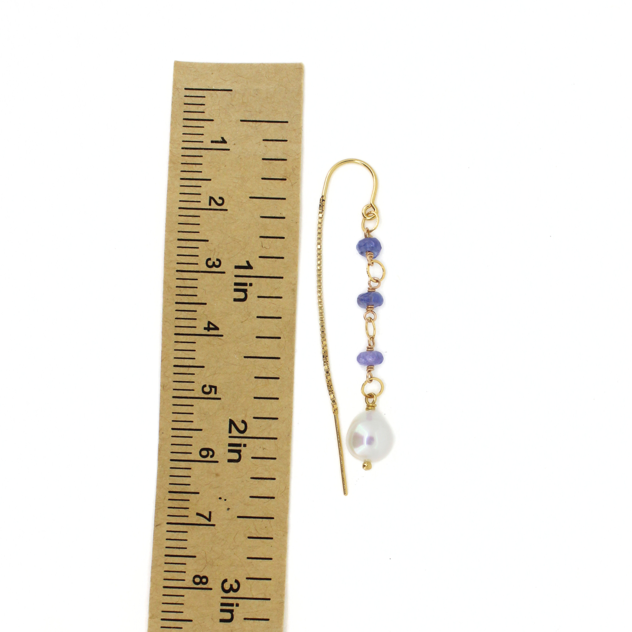 A single gold-toned dangle earring with small purple beads, a white pearl drop, and a chain, shown next to a brown paper ruler for scale on a white background.