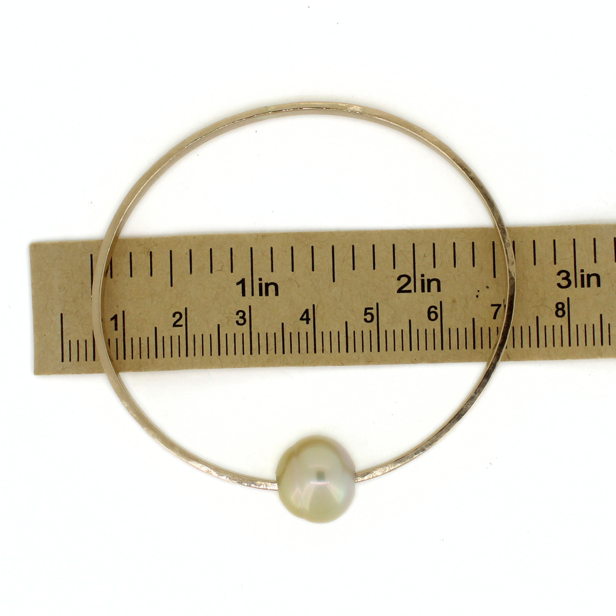 A thick gold oval bangle bracelet with a single irregular gold pearl, shown against a white background with a paper ruler for scale.
