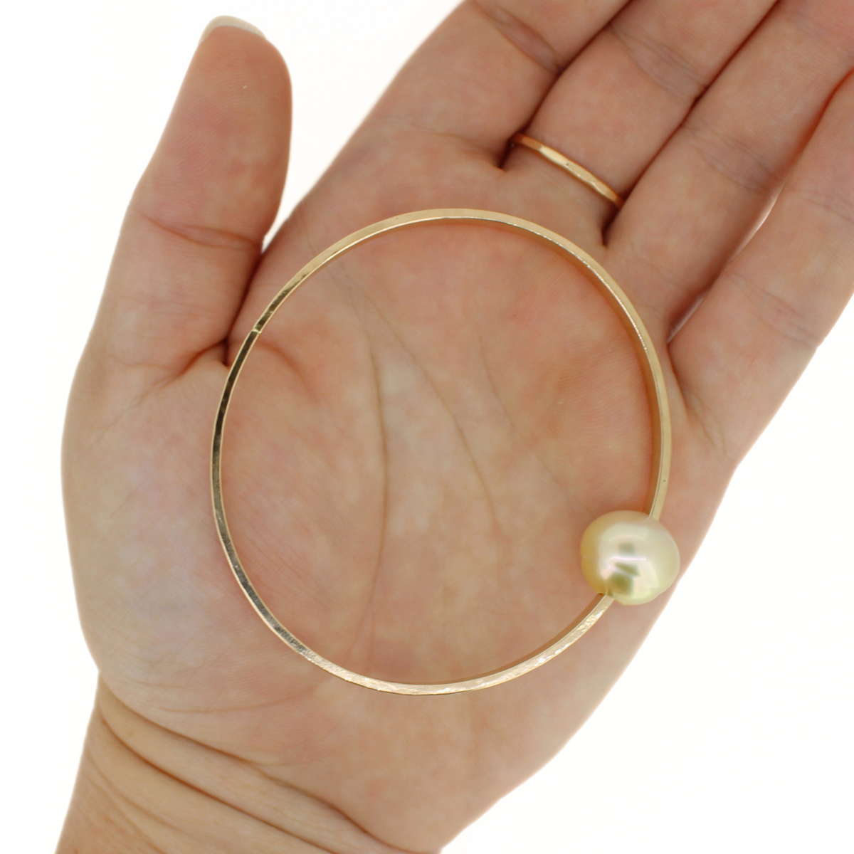 A gold semi-oval bangle bracelet with a single pearl bead, held in the palm of a hand against a white background.