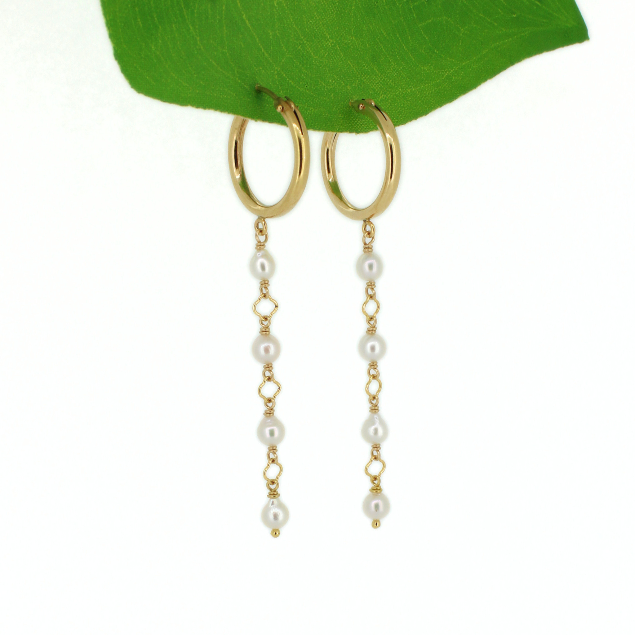 Gold hoop dangle earrings with white pearl bead chains, displayed on a green leaf against a white background.