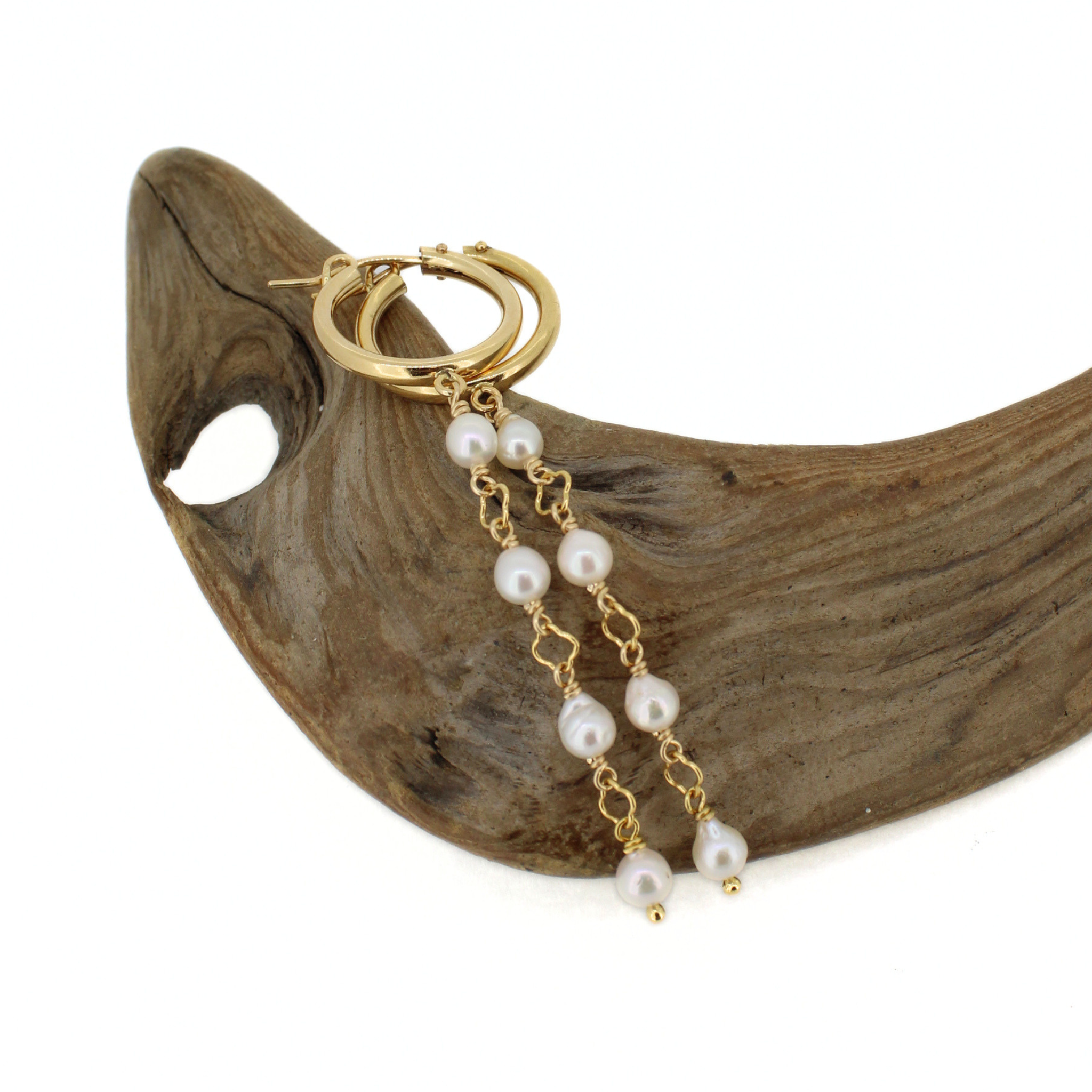 Gold hoop earrings with dangling pearl chains, displayed on a curved piece of natural wood against a white background.