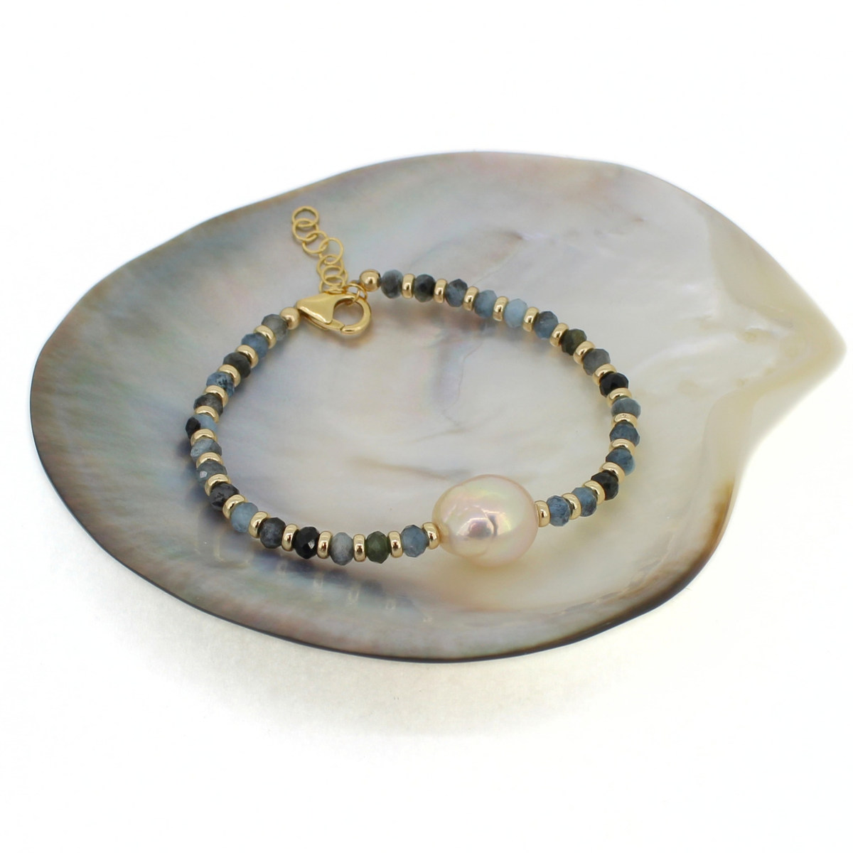 A beaded bracelet with small gold spacer beads, alternating dark and muted blue-gray beads, and a large iridescent pearl, displayed on a large mother-of-pearl shell against a white background.