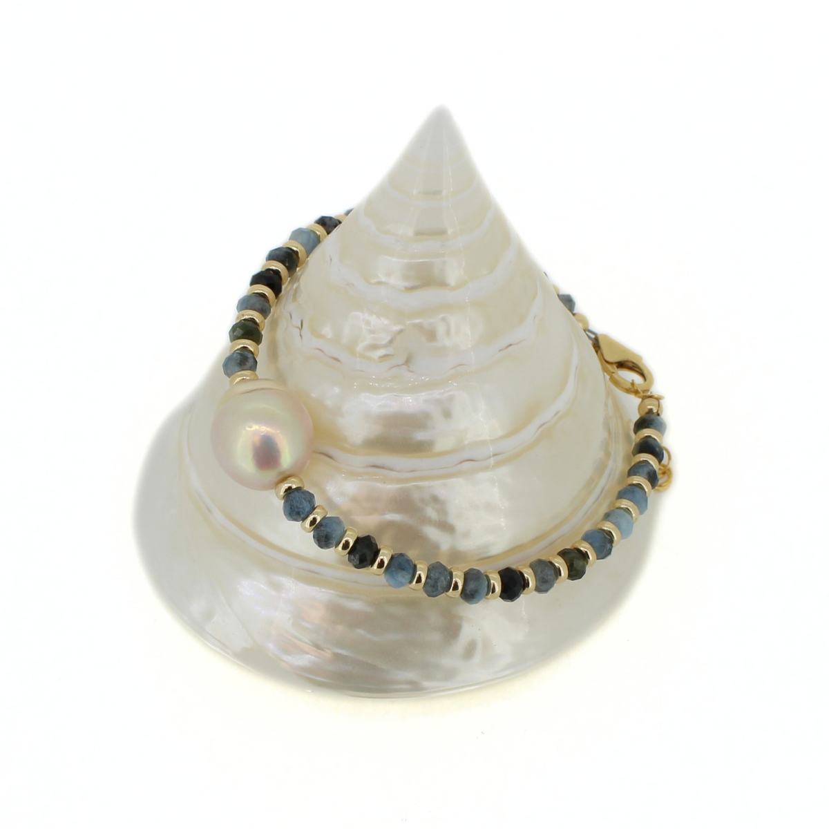 A gold beaded bracelet with dark blue-gray beads, a large iridescent pearl, and a gold clasp, displayed on a white mother-of-pearl seashell stand against a white background.