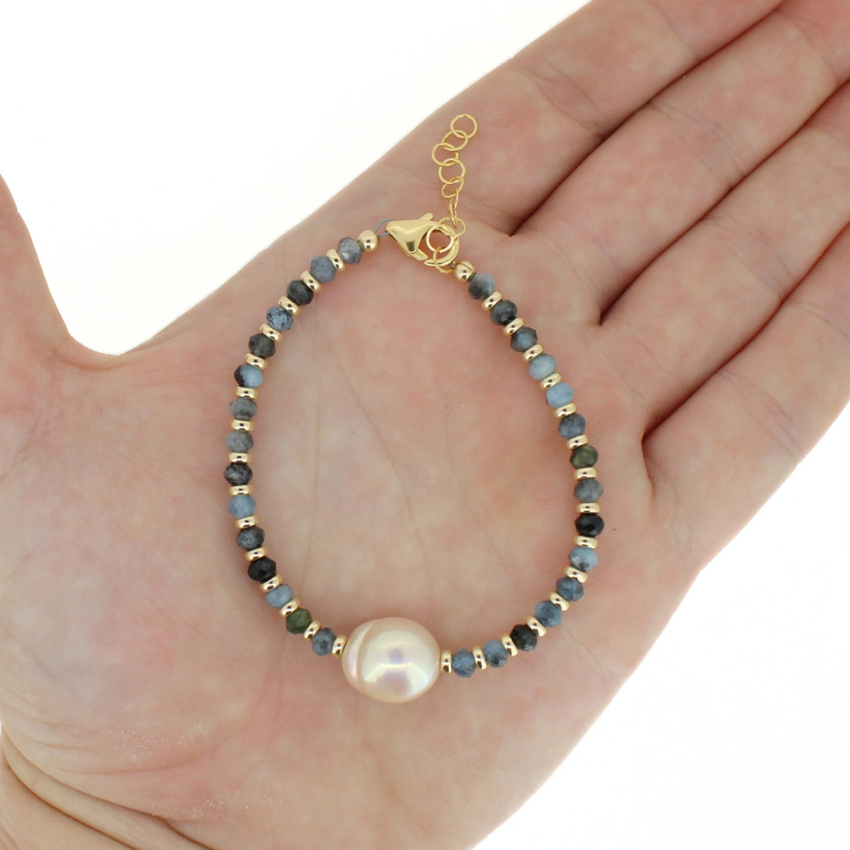 A delicate beaded bracelet with a large white pearl, displayed on an open hand. The bracelet features small faceted beads in shades of blue, gray, and green, separated by gold spacer beads, and finished with a gold clasp.
