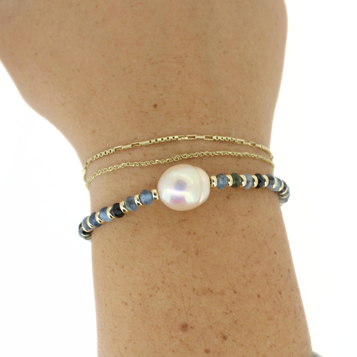A close-up of a person's wrist wearing a delicate double-strand bracelet stack. The top layer is a fine gold chain, and the bottom layer features a beaded bracelet with a large, lustrous white freshwater pearl in the center, flanked by small gold beads and blue-green stones.