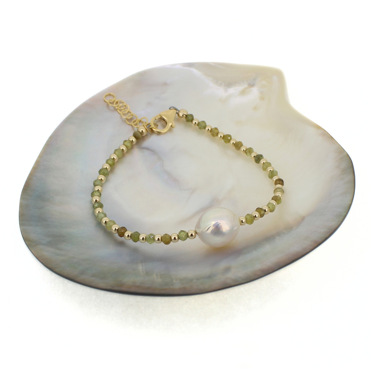 A delicate gold bracelet with small green and gold beads and a large white pearl, displayed on a white mother-of-pearl clam shell.