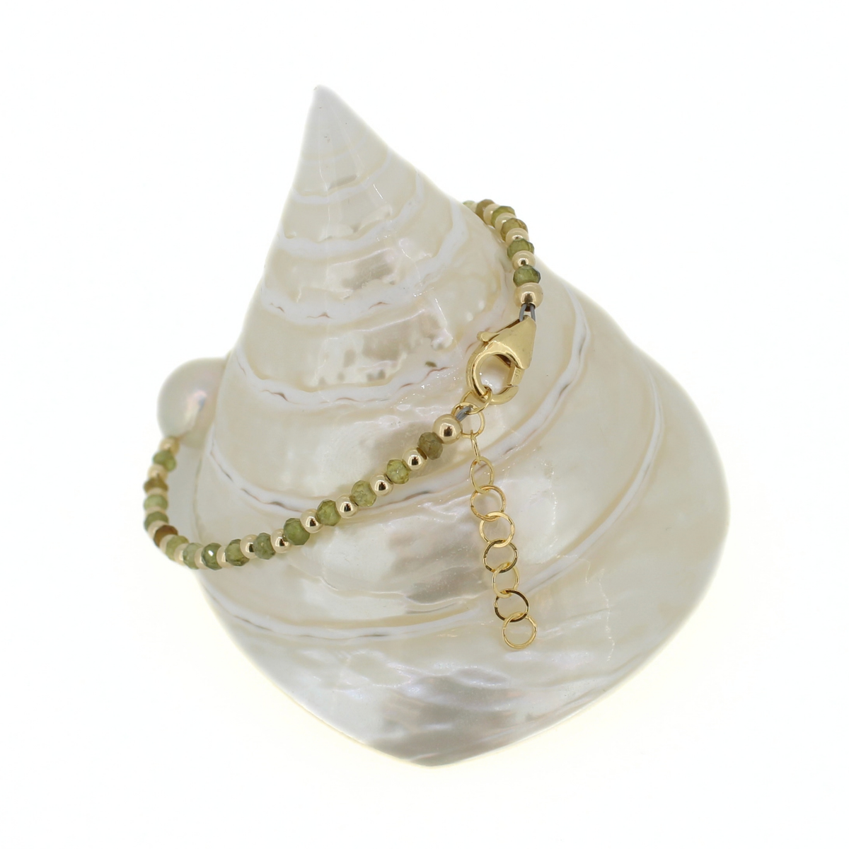 A delicate beaded bracelet with olive green beads and gold metal hardware, displayed on a white mother-of-pearl seashell against a white background.