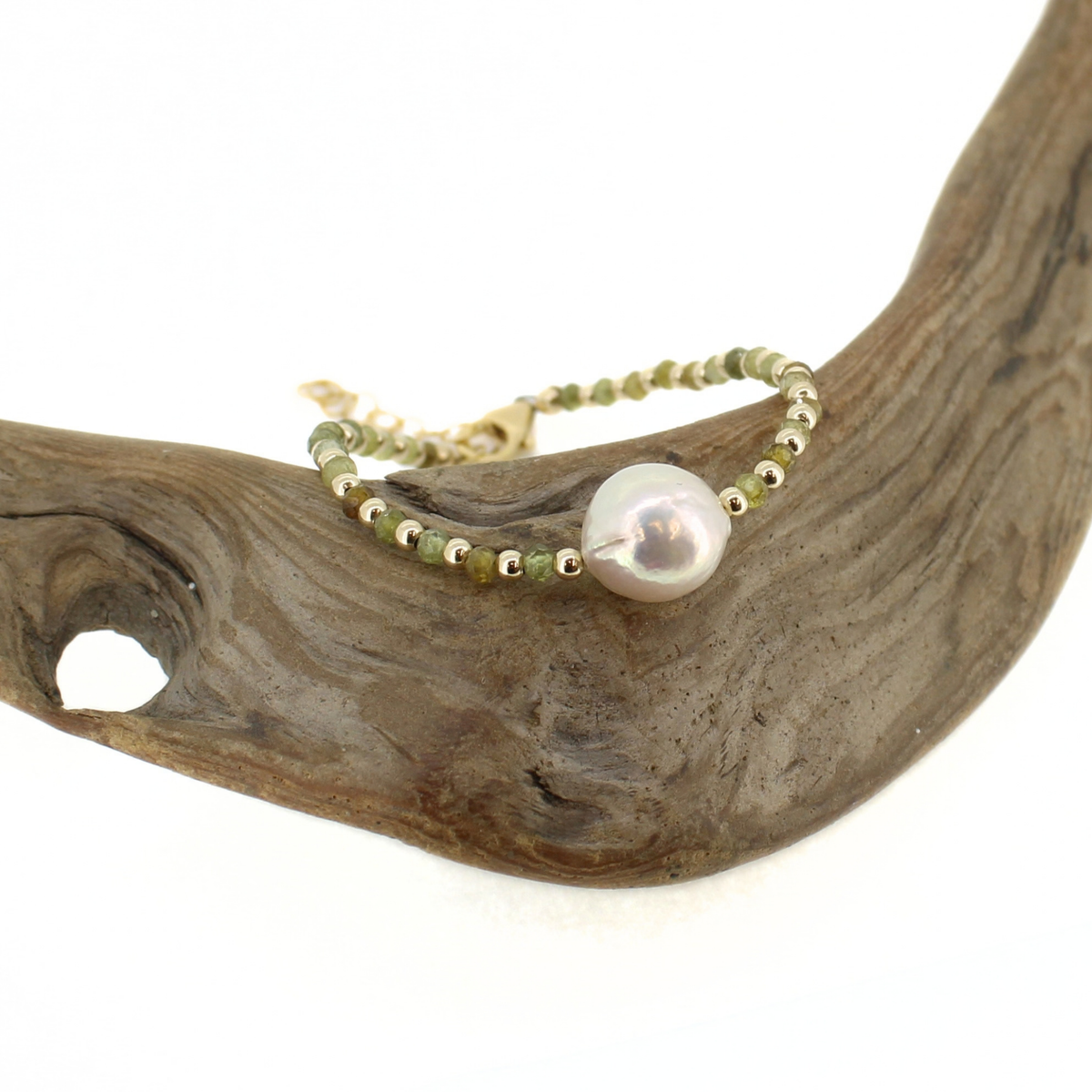 A delicate beaded bracelet with a large white pearl and small metallic beads, displayed on a curved piece of driftwood against a white background.