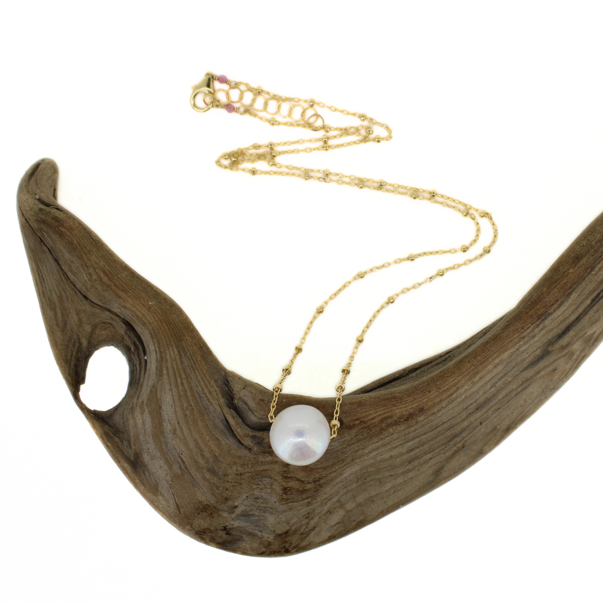 A dainty gold satellite chain featuring a white fresh water pearl pendant, displayed on a piece of driftwood.