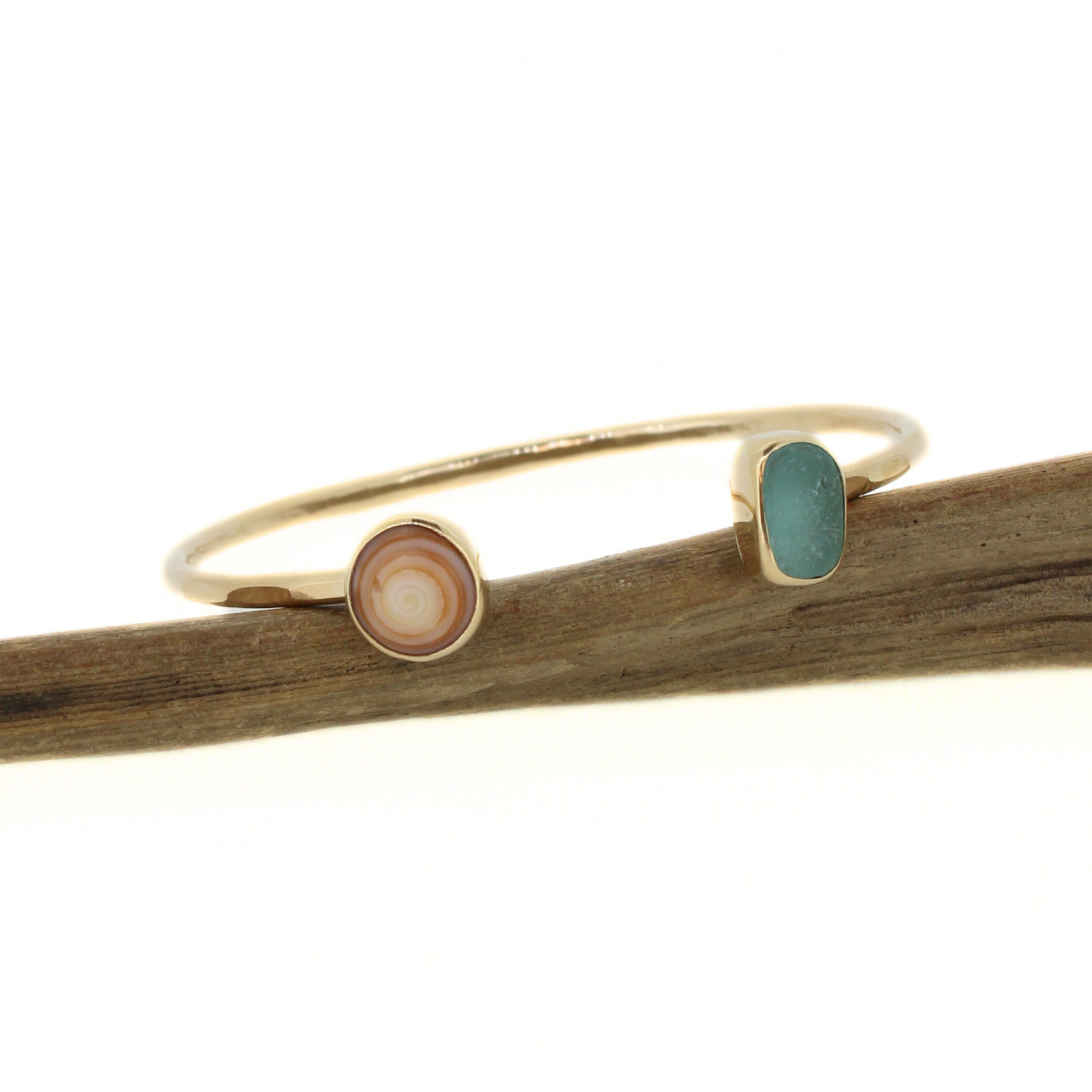 A slim gold cuff bracelet with two round bezel-set stones, one a brown puka shell and one deep blue sea glass, displayed on a piece of wood against a white background. Handmade in Hawaii by Brianne & Co.