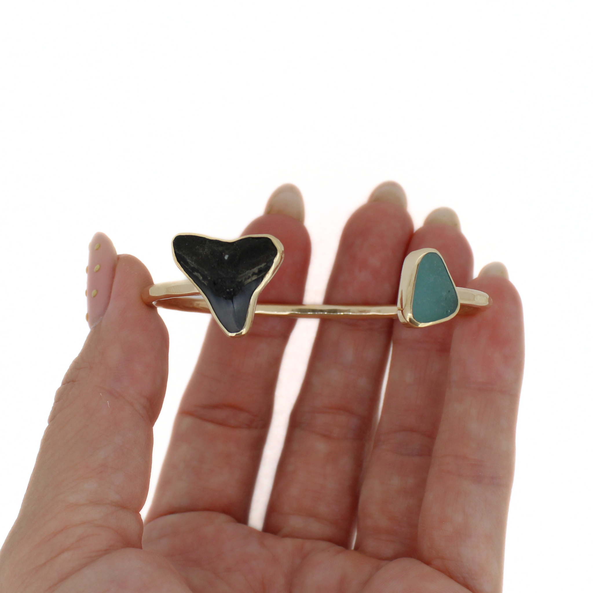 Gold cuff bracelet held in hand with black shark tooth and green sea-glass triangle tips