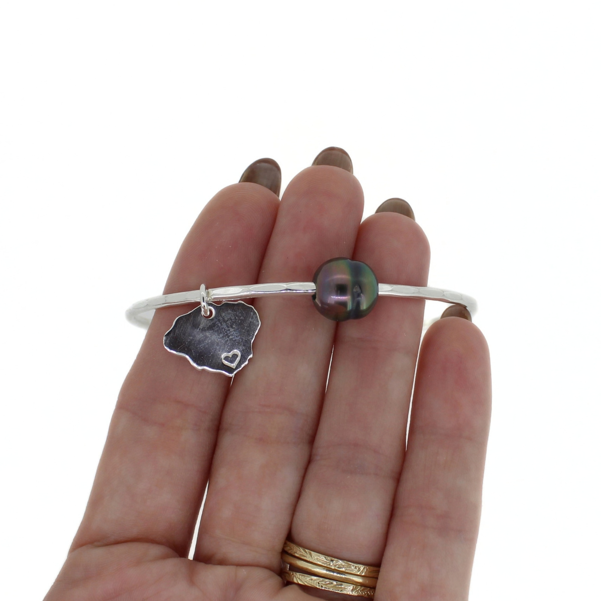 Sterling silver bangle bracelet with a dark Tahitian pearl and a Kauai island charm shown on a persons hand for size reference 