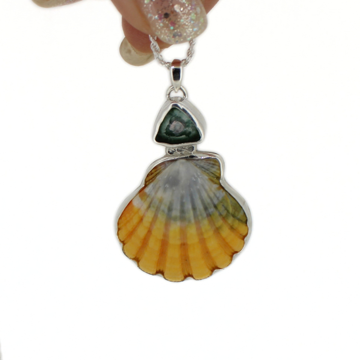 A silver pendant necklace featuring a sunrise shell and tourmaline gemstone, suspended from a silver chain and held by a hand with glittery nail polish against a white background.