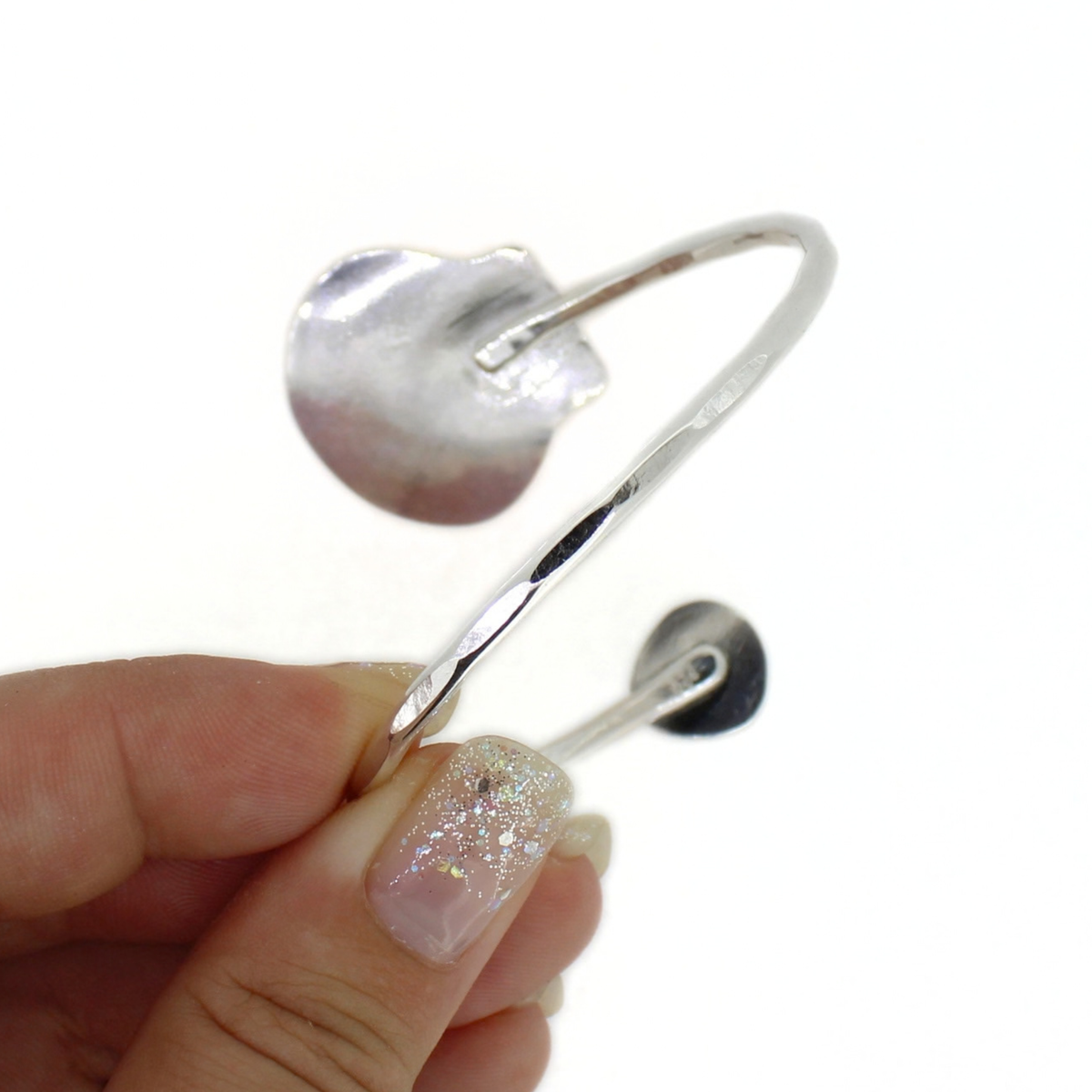 A hand with glittery nail polish holds a polished silver open cuff bracelet against a plain white background.
