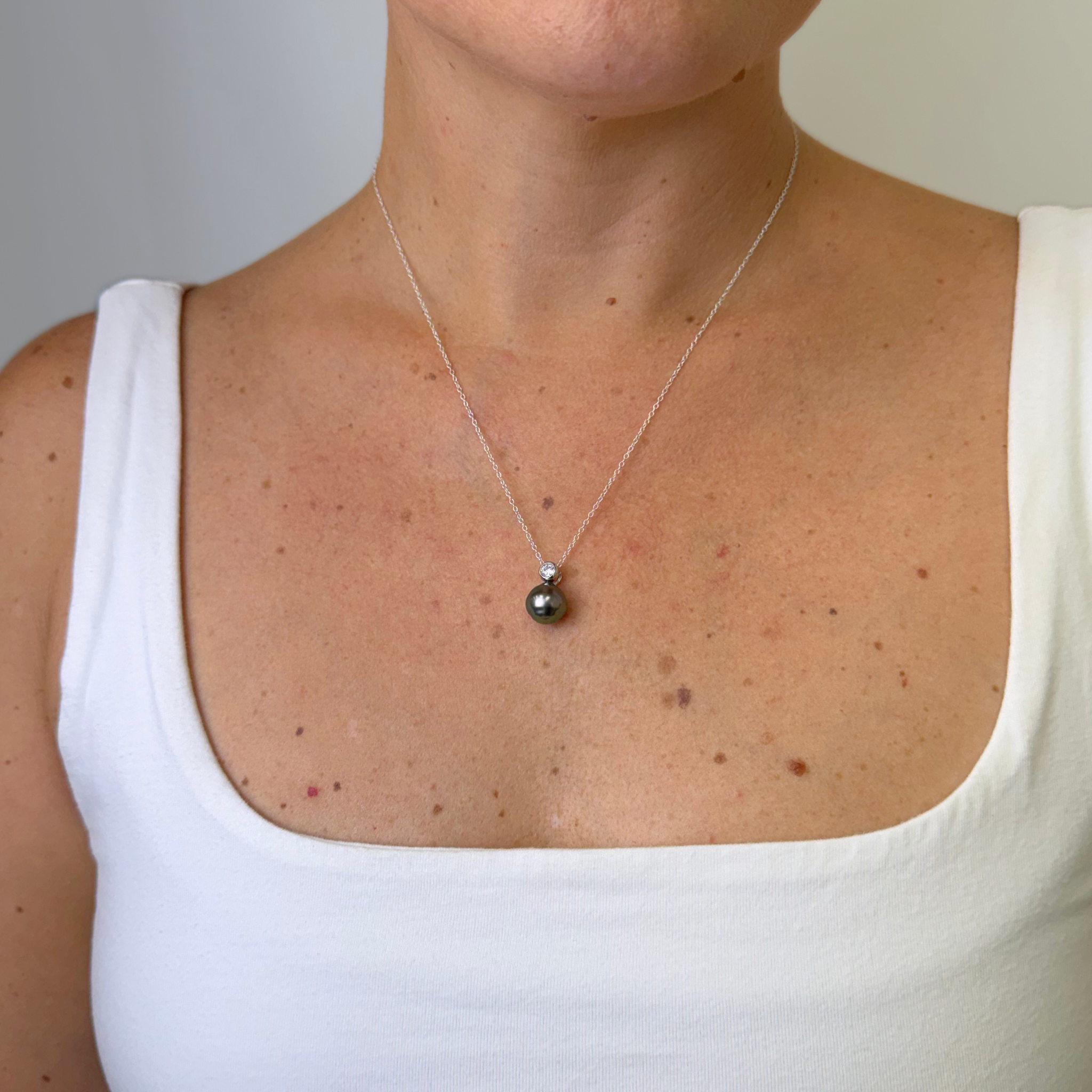 A close-up of a person wearing a delicate silver chain necklace with a small dark gray pearl pendant, styled with a white tank top.