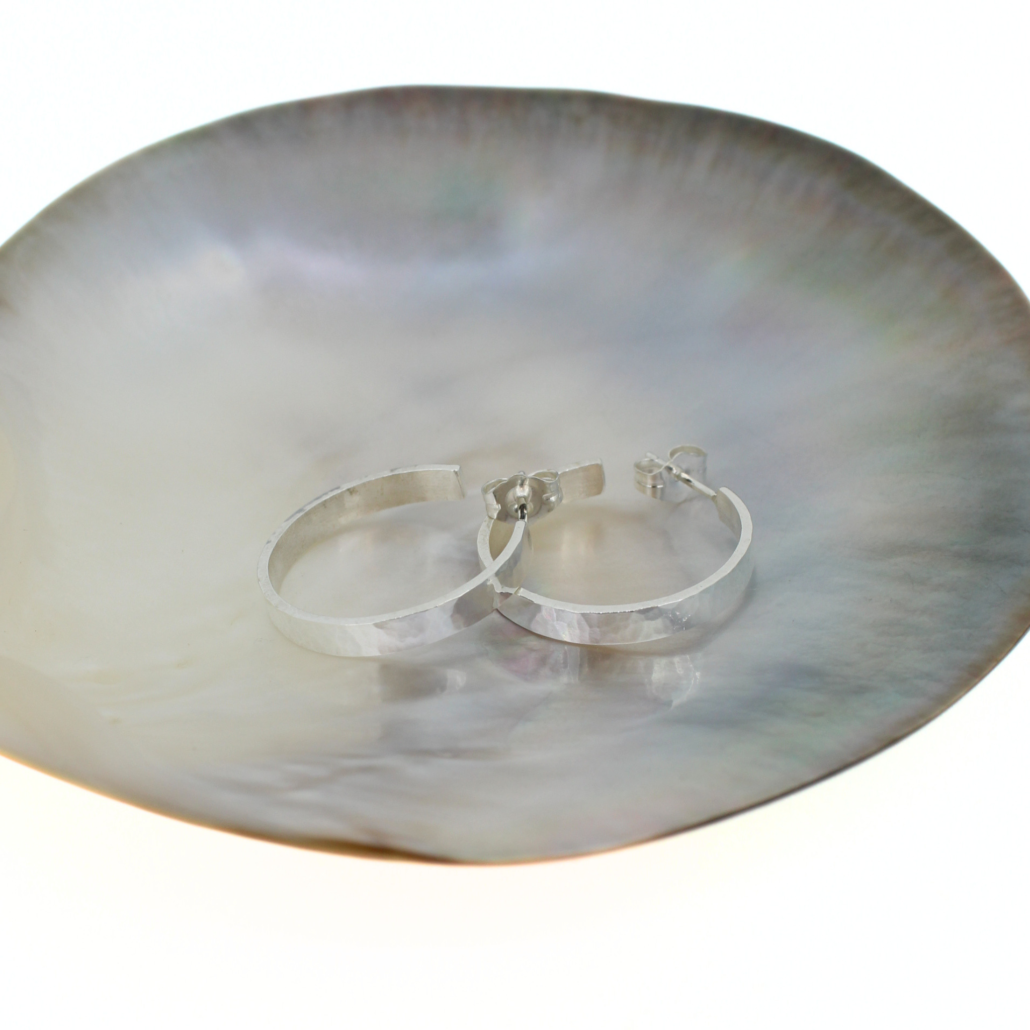 A pair of polished silver hoop earrings resting on a large, iridescent mother-of-pearl dish against a white background. Handmade by Brianne & Co. in Kauai, Hawaii
