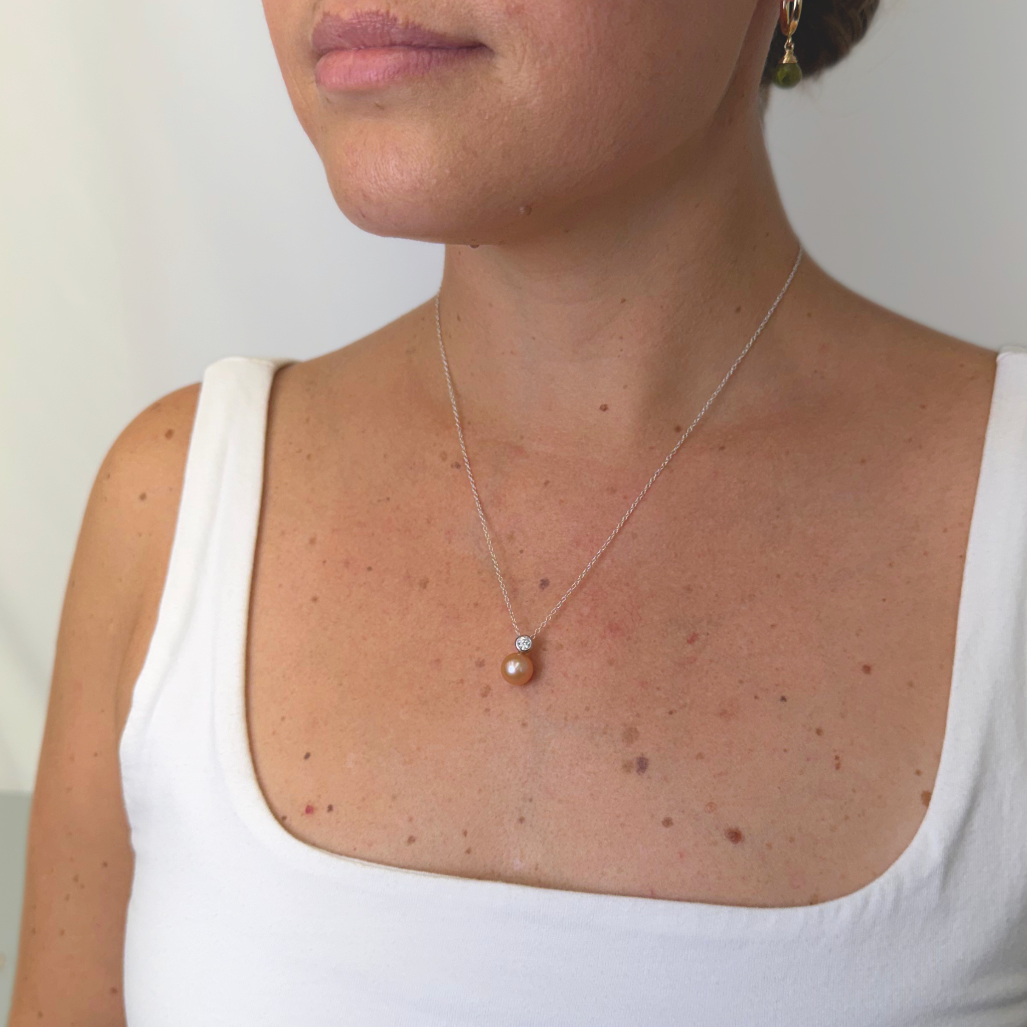 A close-up of a woman wearing a delicate silver chain necklace with a small white crystal and a single peach-colored pearl pendant, paired with a white tank top.